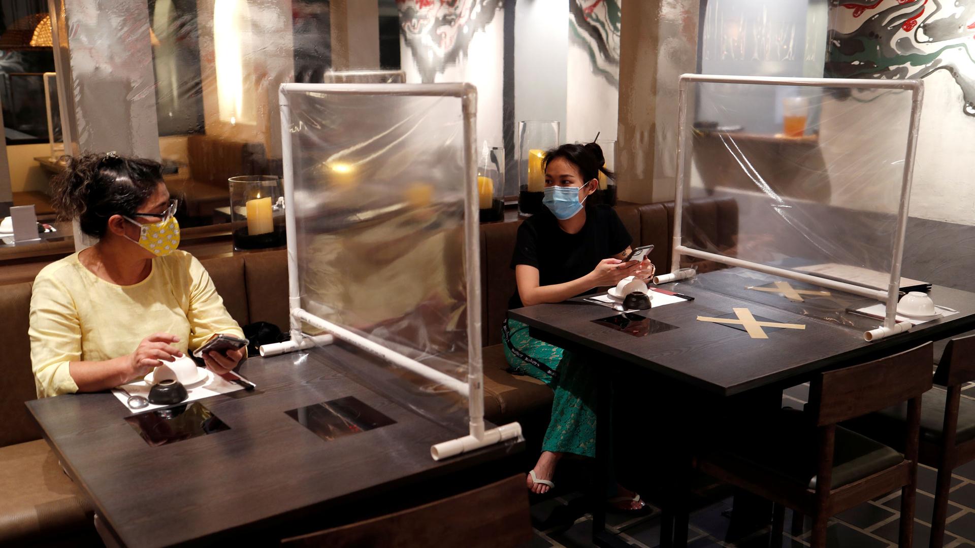 Two women are shown sitting at separate tables, wearing protective face masks with a plastic screen in-between them.