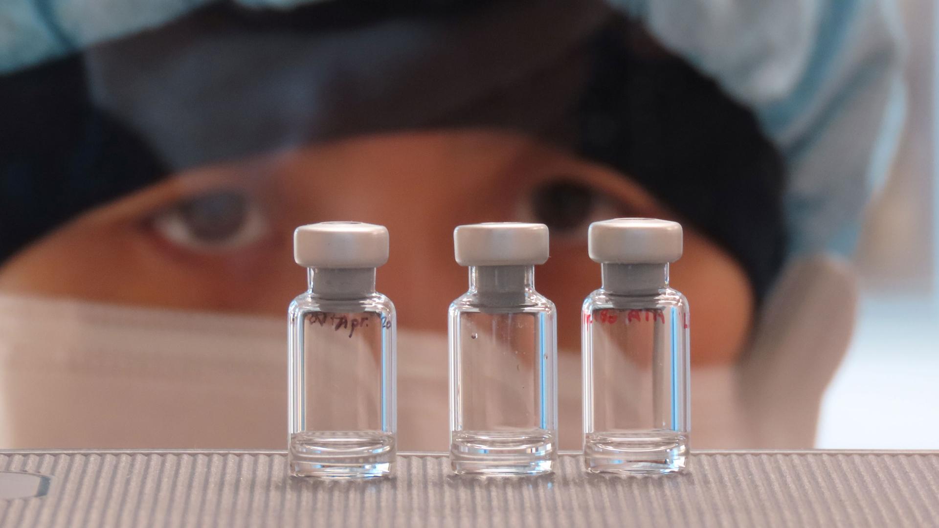 Three small, clear glass vials are shown with the eyes of a scientist looking at them in the background.