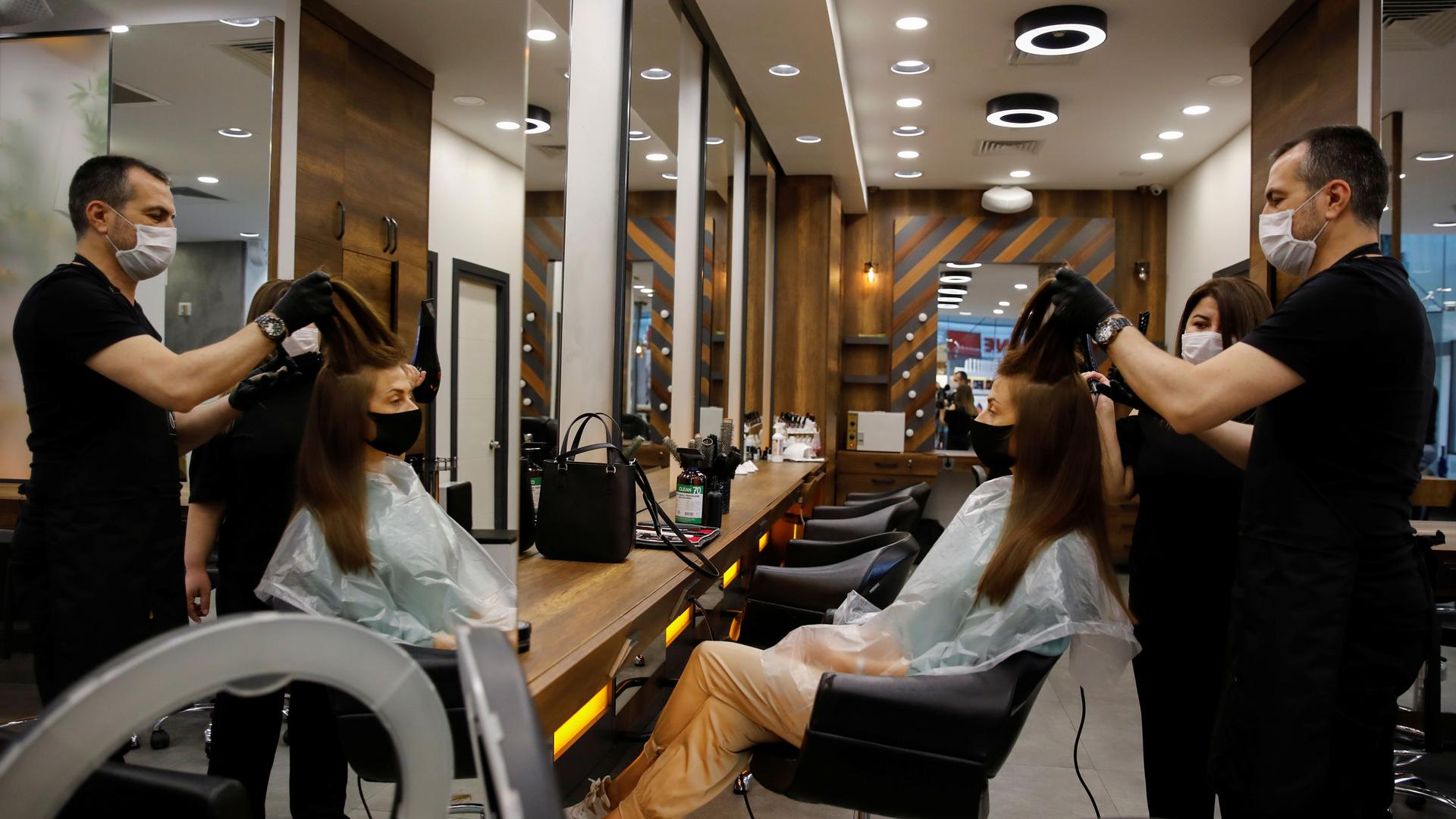 A hairdresser is shown standing behind a customer who is sitting in a chair while he holds her hair up in the air.