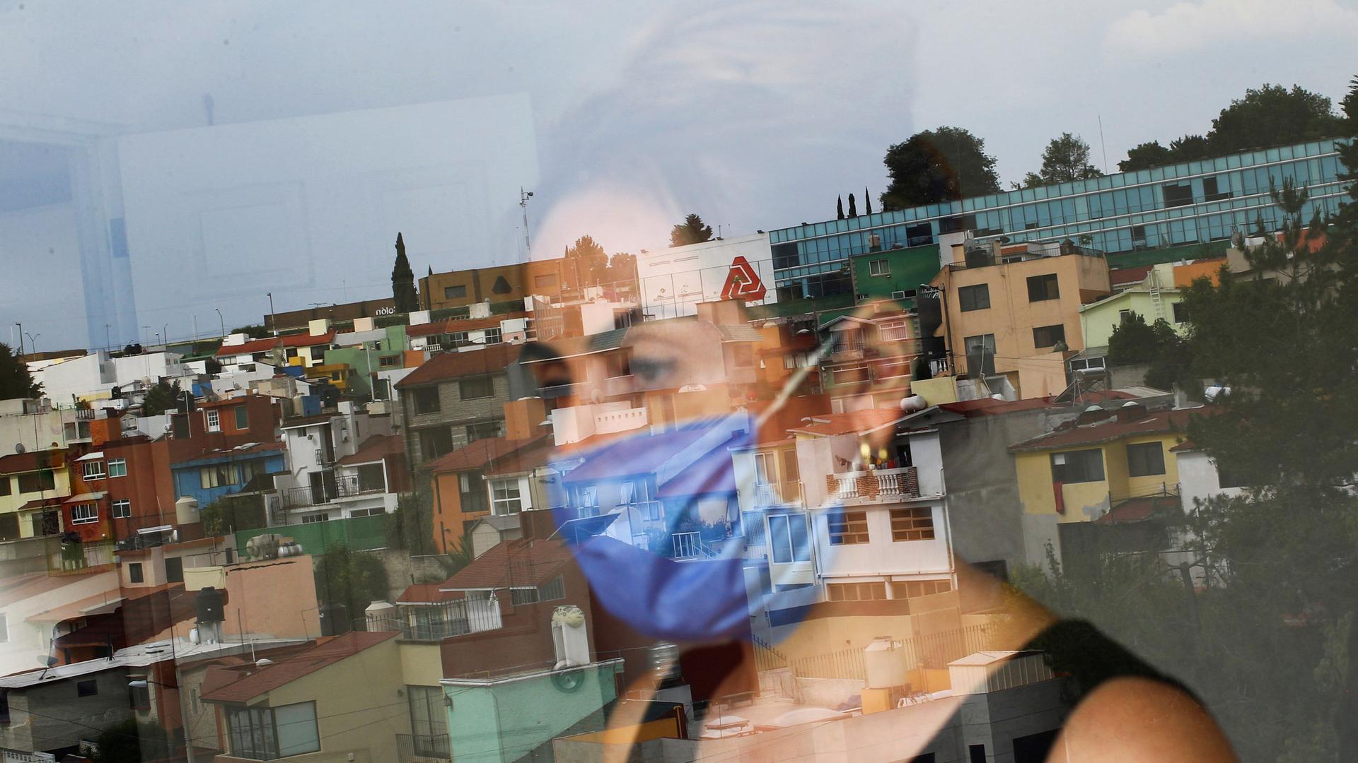 A woman is shown through a clear glass window with the reflection of a Mexico City neighborhood.
