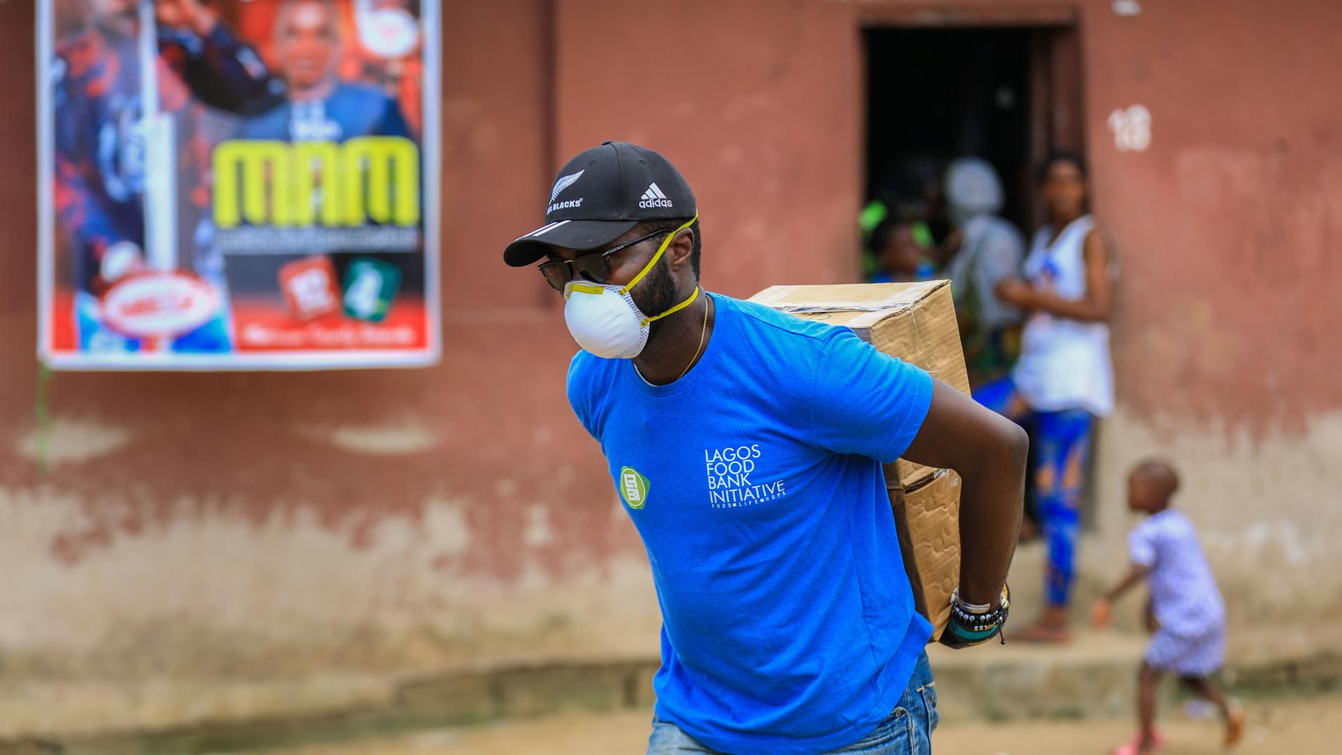 A man is shown wearing a blue shirt with "Lagos Food Bank" written on it while carrying a cardboard box on his back.