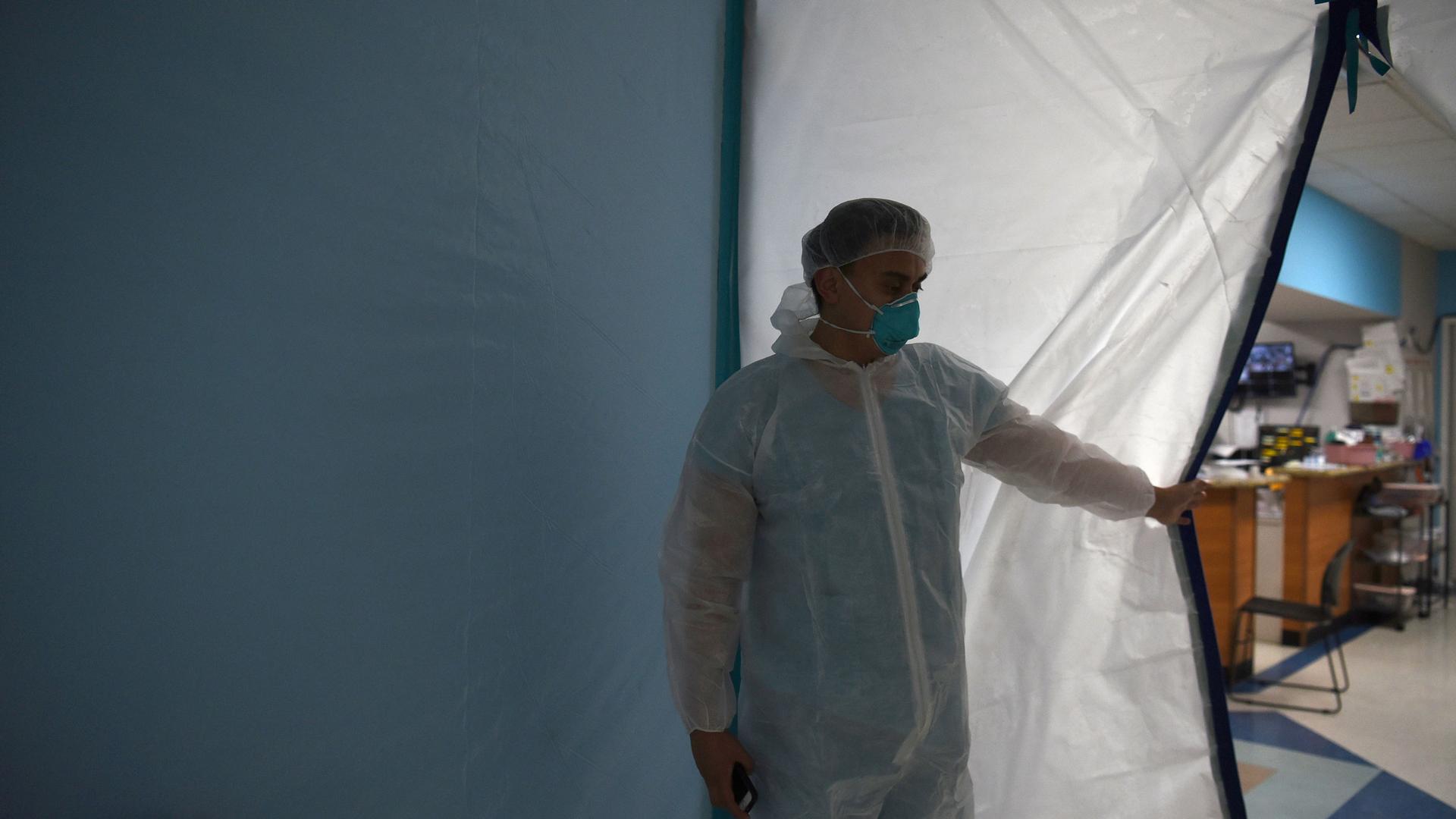 A man is shown wearing medical protective clothing and a face mask while holding open a plastic door.