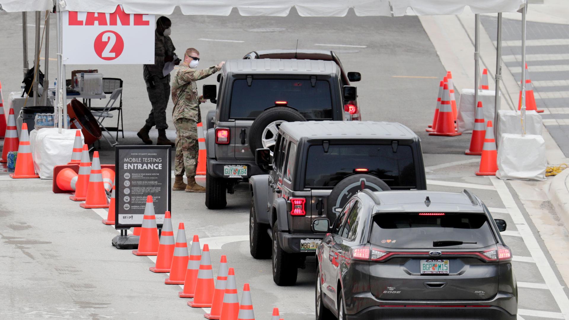 A line of vehicles are shown next to a row of orange traffic cones leading to a National Guard official at a mobile coronavirus testing facility.