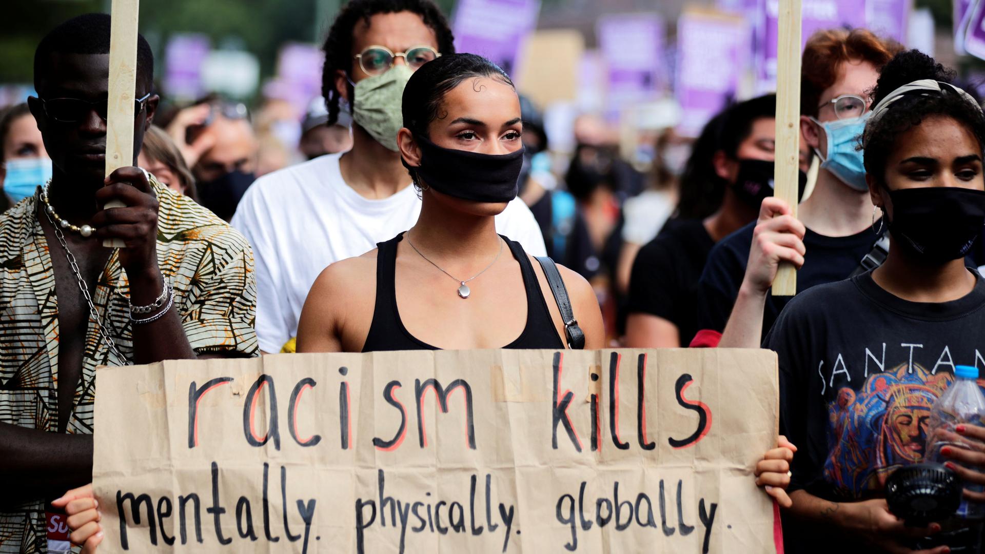 A large crowd of people are shown with a women featured in the center of the photograph holding a sign that reads, racism kills, mentally. physically. globally.