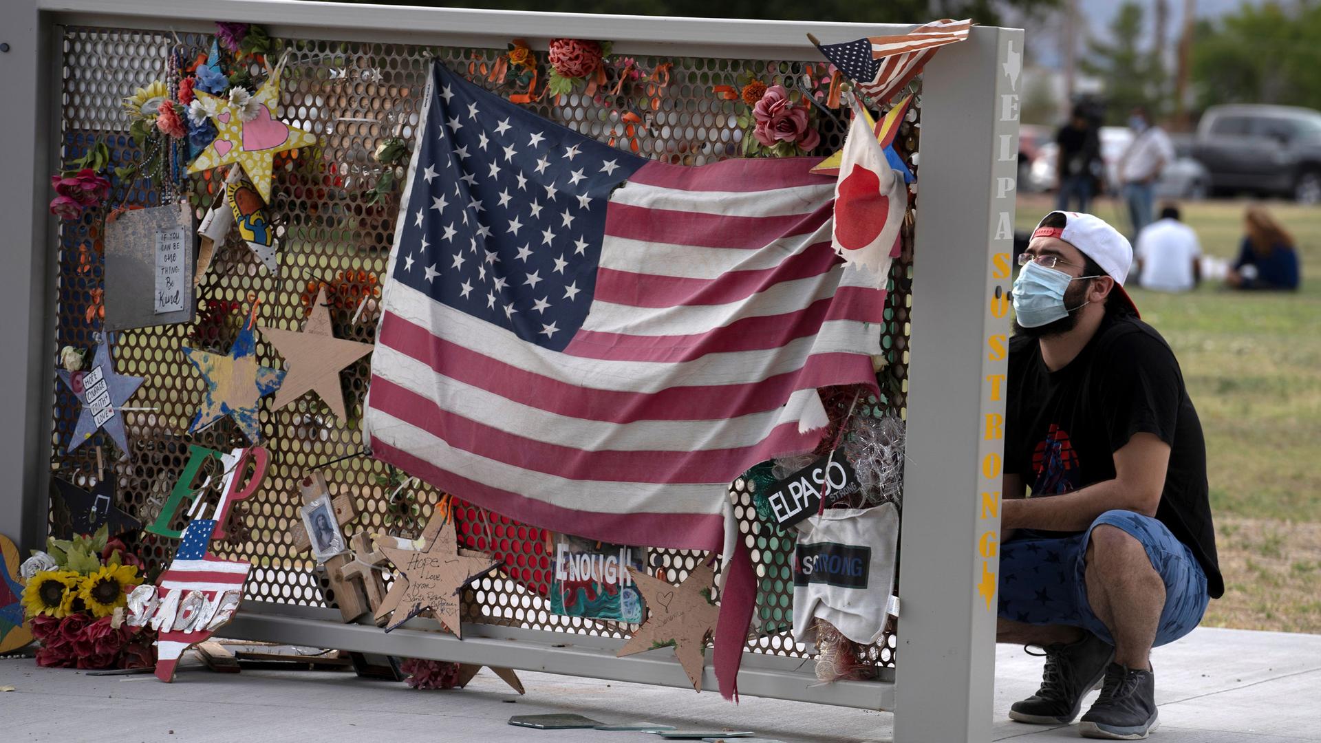 A man is shown kneeling next to a memorial that has stars and an American flag pinned on it.