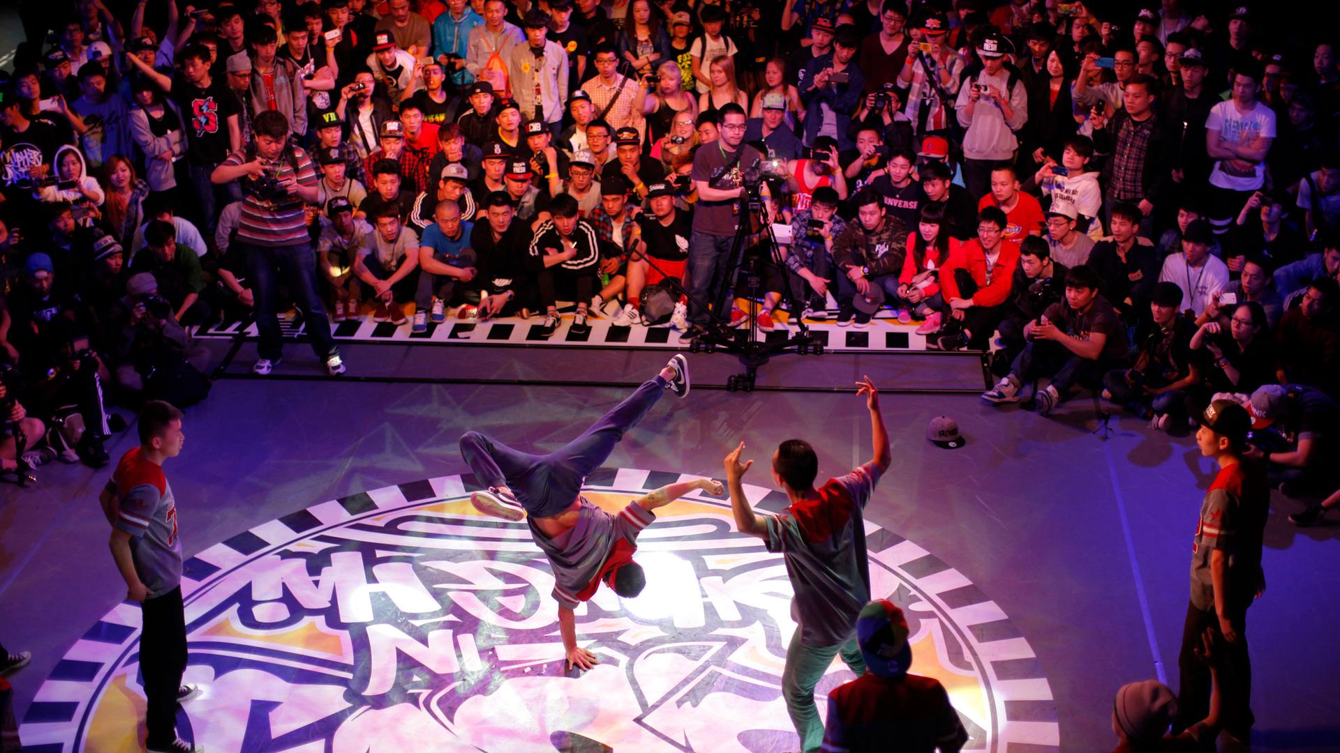 Several breakdancers perform on stage with a large crowd watching.