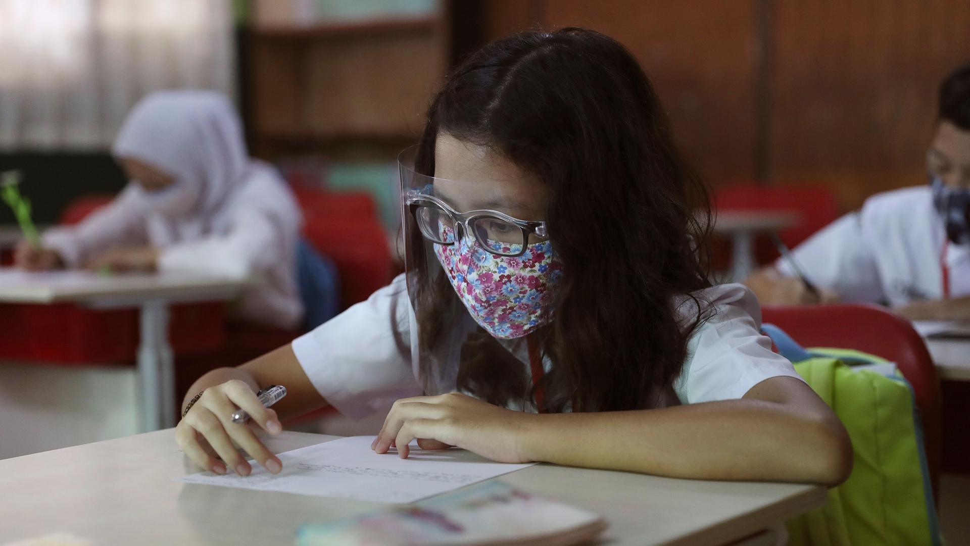 A young girl is shown sitting at a classroom desk wearing a protective face mask and plastic face guard while holding a pen.