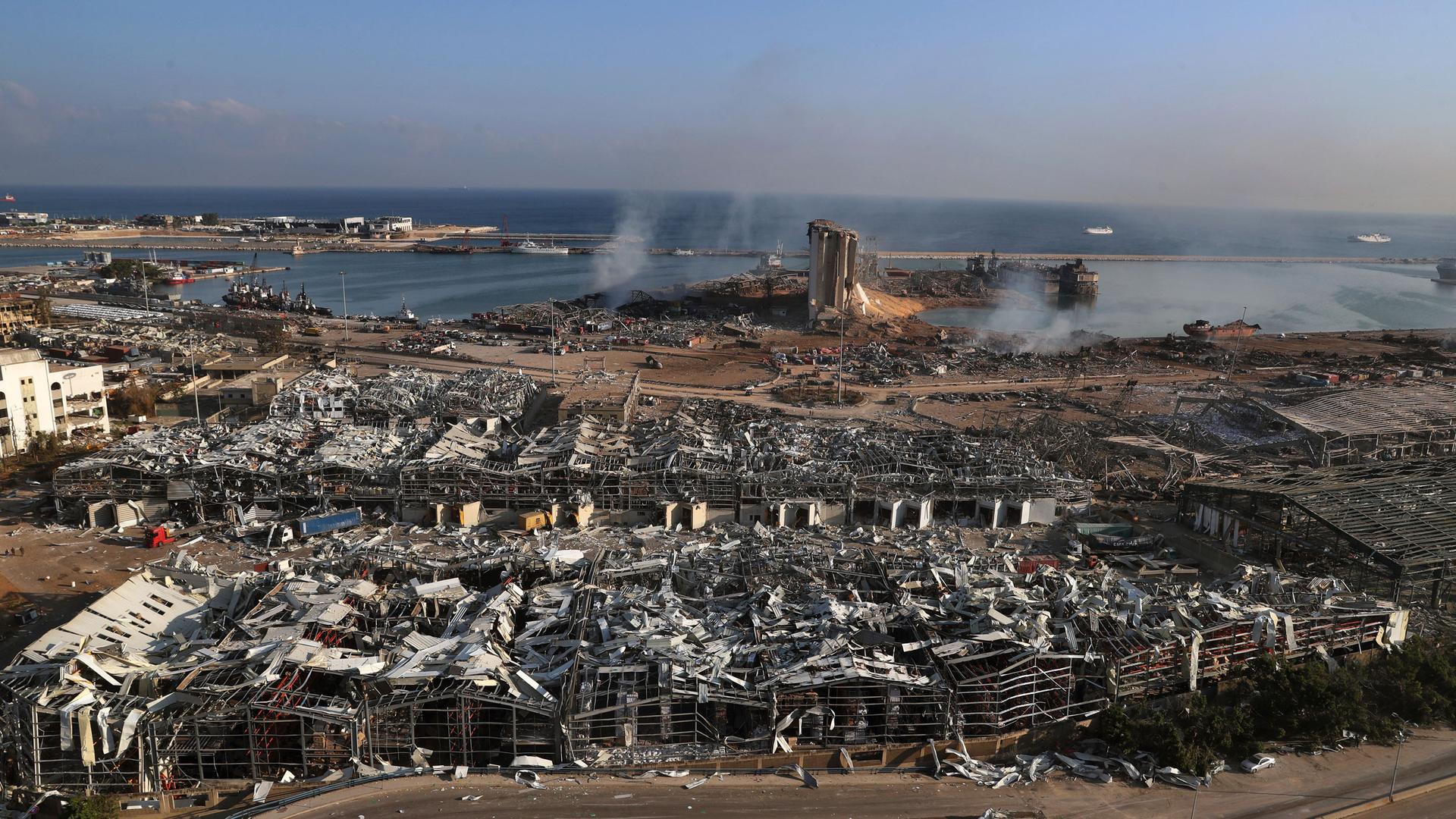 A photo showing Beirut's port left in complete rubble with smoke rising from parts and the Mediterranean Sea in the distance.