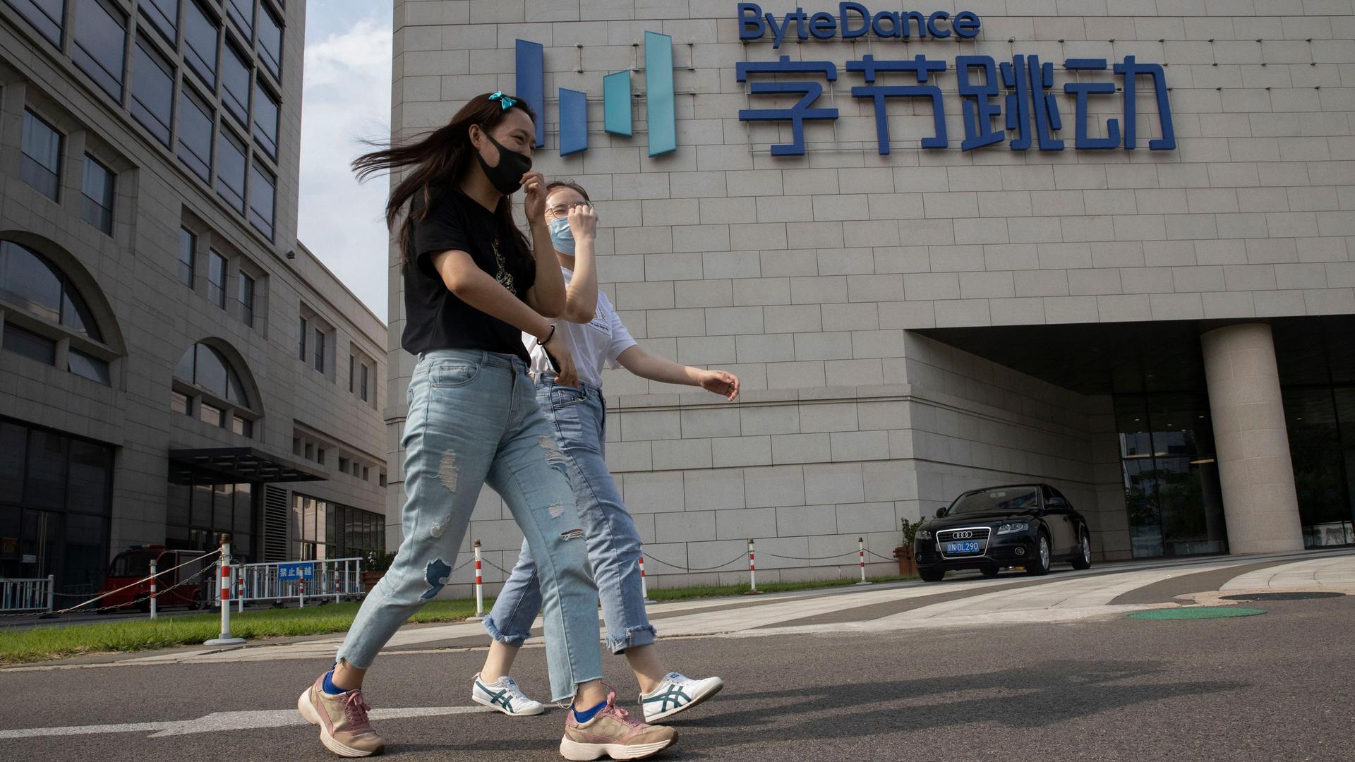 Two women are shown wearing jeans and face masks while walking past the ByteDance headquarters with large English and Chinese writing on the side.