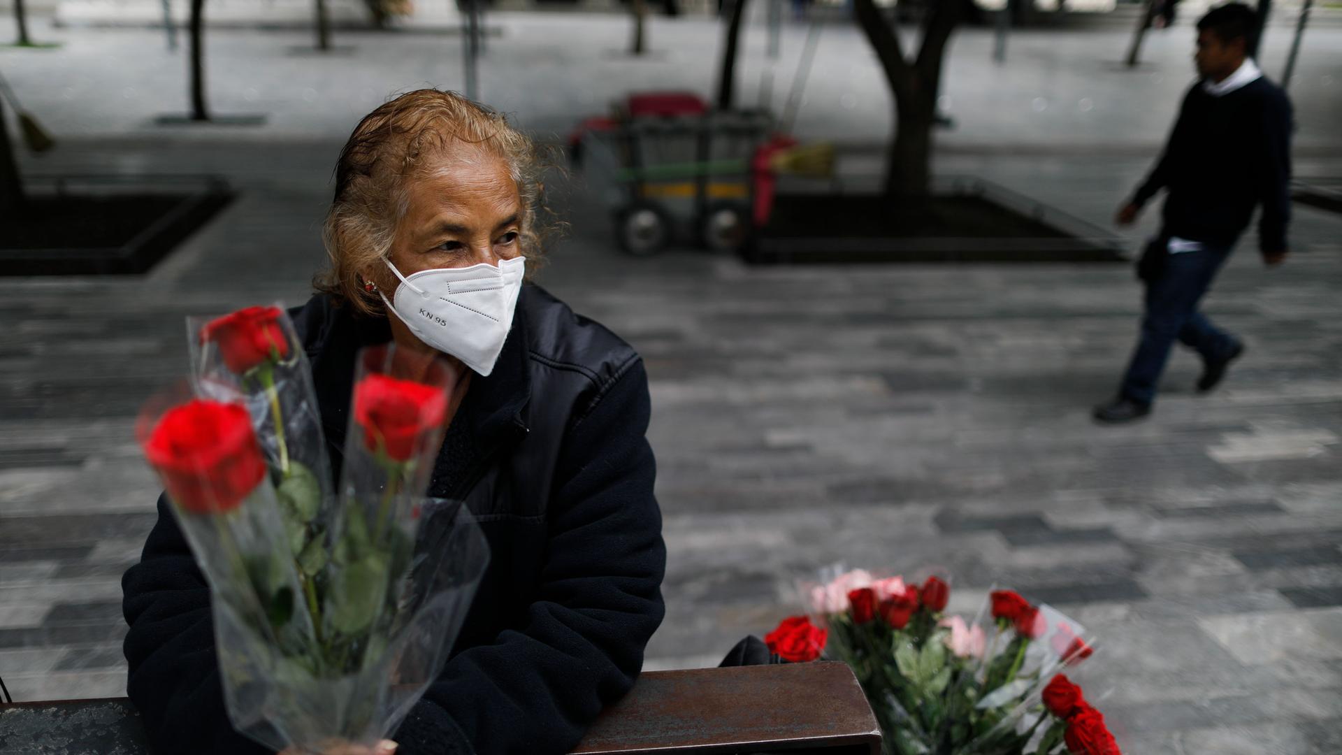 A woman is shown sitting on a metal bench and holding three red roses while wearing a protective face mask.