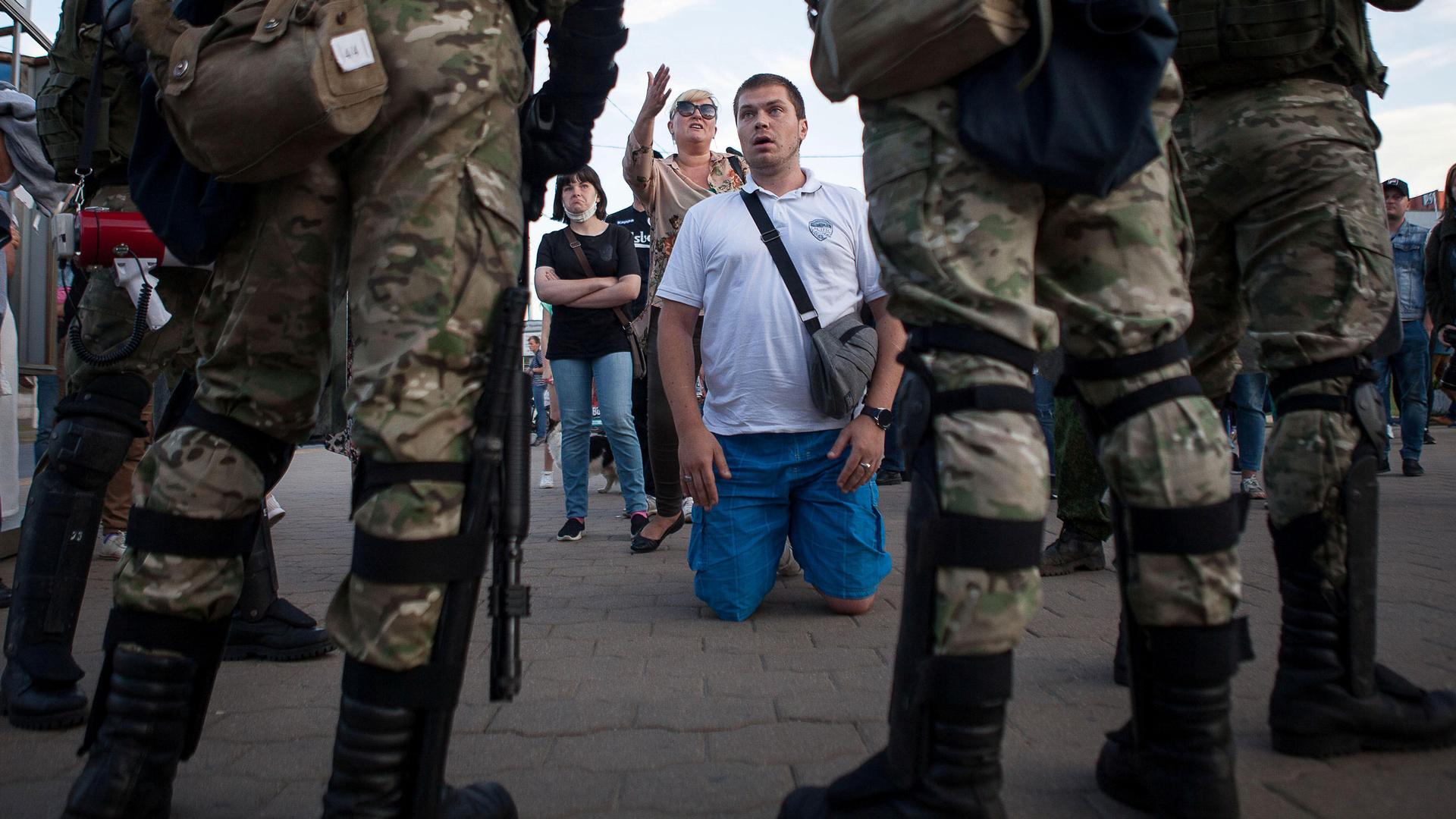 A man is shown kneeling on the ground in front of several security officers dressed in camoflage and carrying weapons.