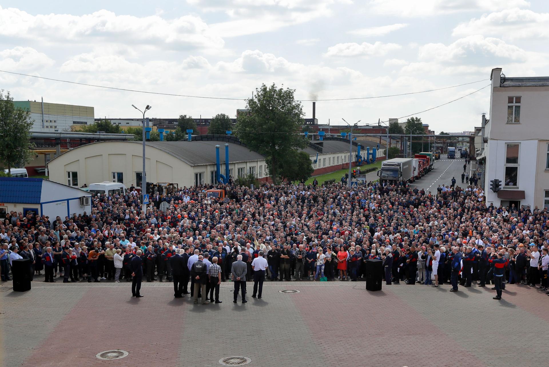 A large crowd of people are shown in the street with a factory in the background.