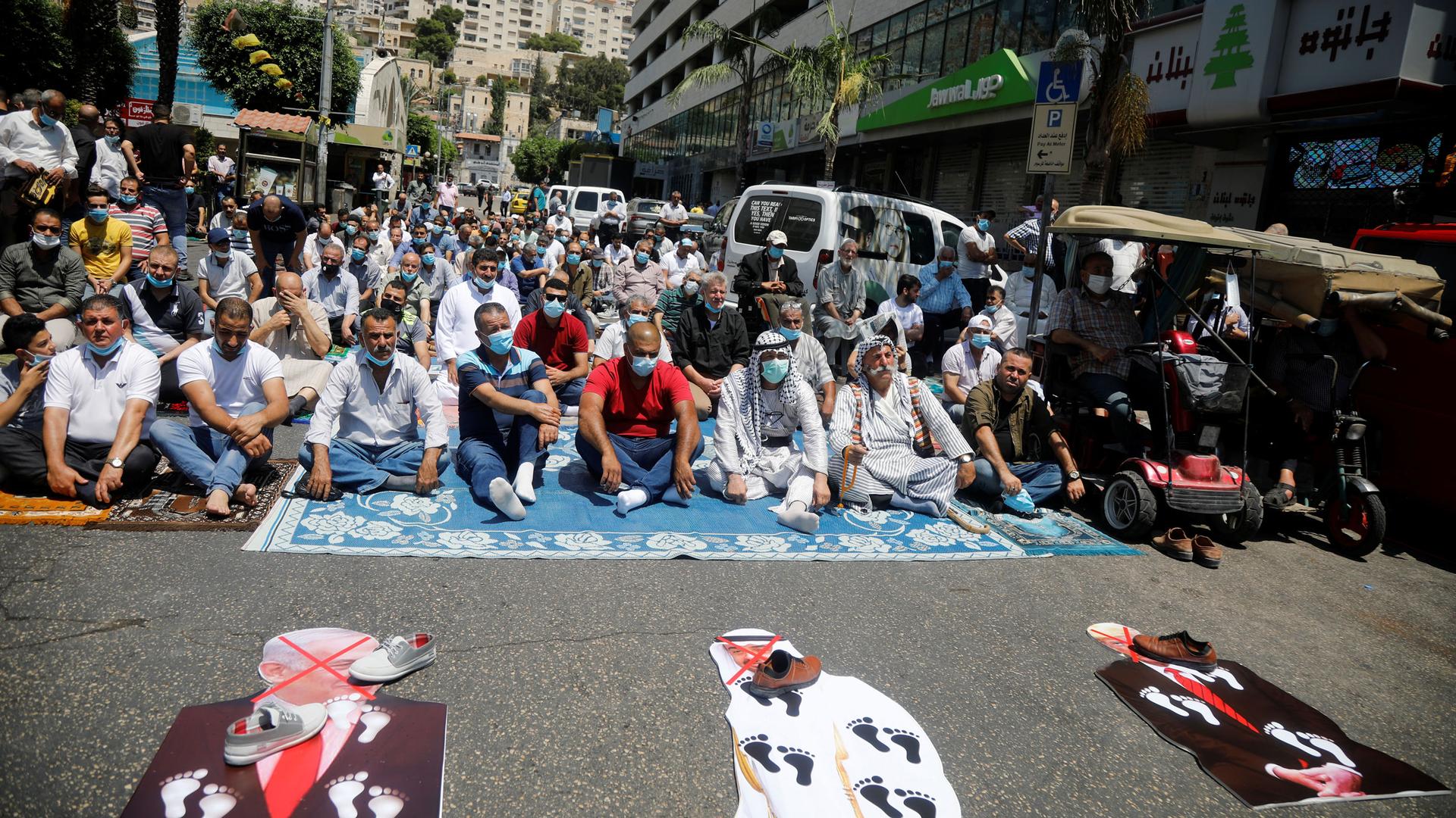 A large group of men are shown sitting in a street with cardboard cutouts of President Donald Trump and Abu Dhabi Crown Prince Mohammed bin Zayed al-Nahyan and Israeli Prime Minister Benjamin Netanyahu lay on the ground in front of them with shoes placed.