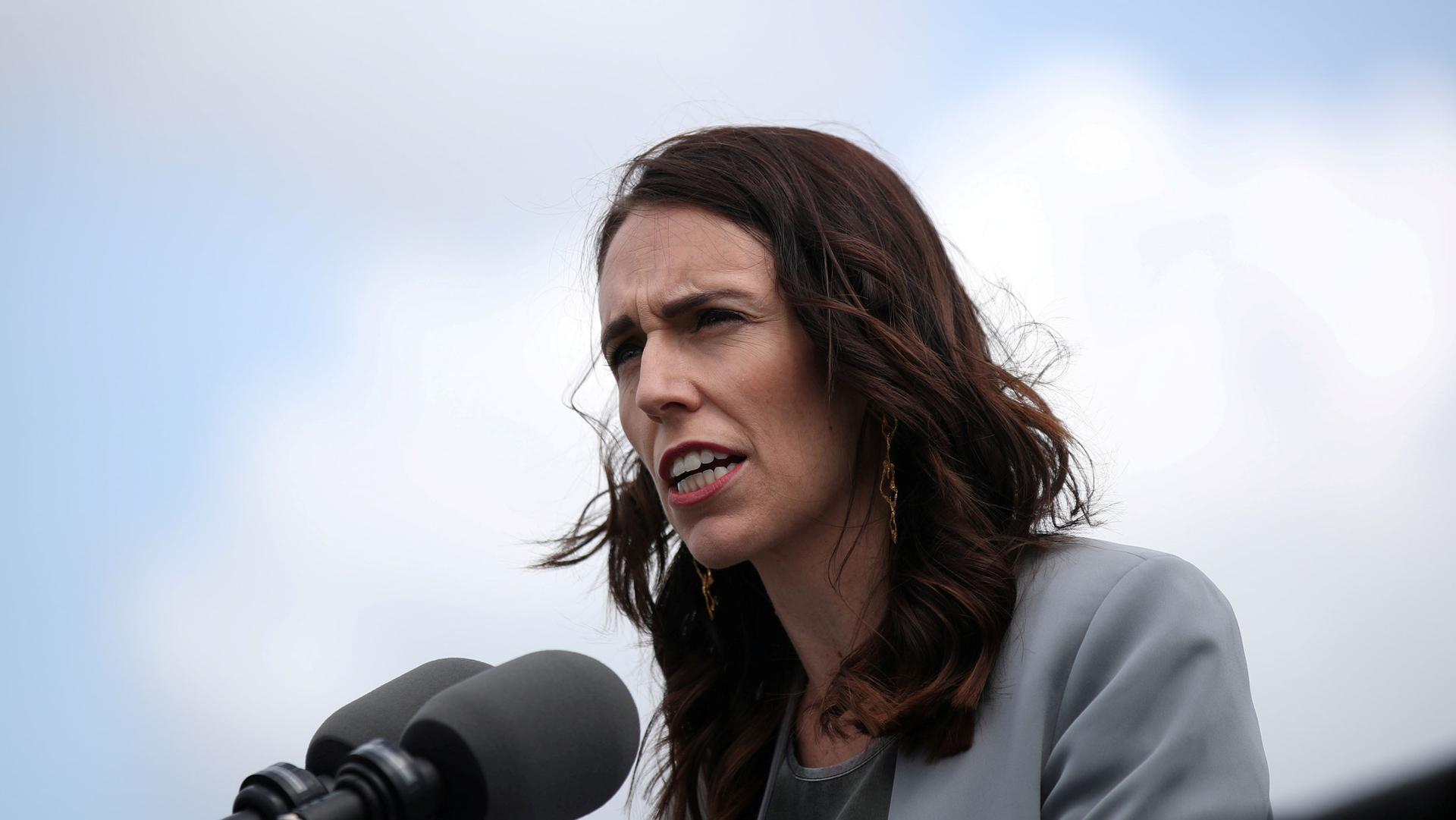 New Zealand Prime Minister Jacinda Ardern is shown outside wearing a gray jacket and speaking into two microphones.