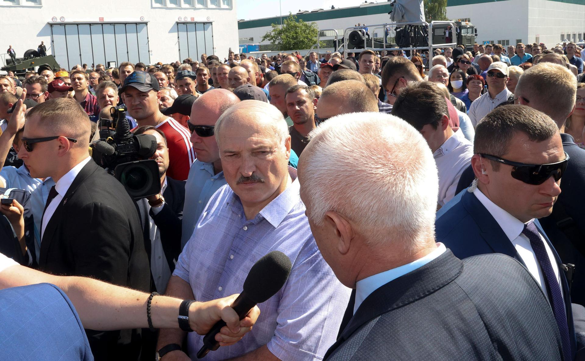 Belarusian President Alexander Lukashenko is shown wearing a blue button down shirt and standing in a large crowd with bodyguards surrounding him.