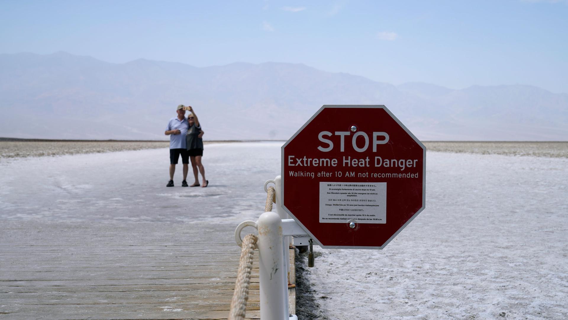 A sign the shape of a red stop sign warns of extreme heat danger at Badwater Basin with a couple taking a selfie in the distance.