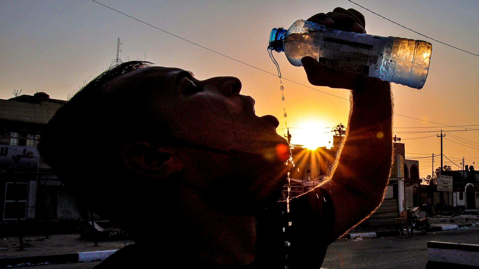 A man is shown in shadow drinking from a water bottle with the sun setting behind him.