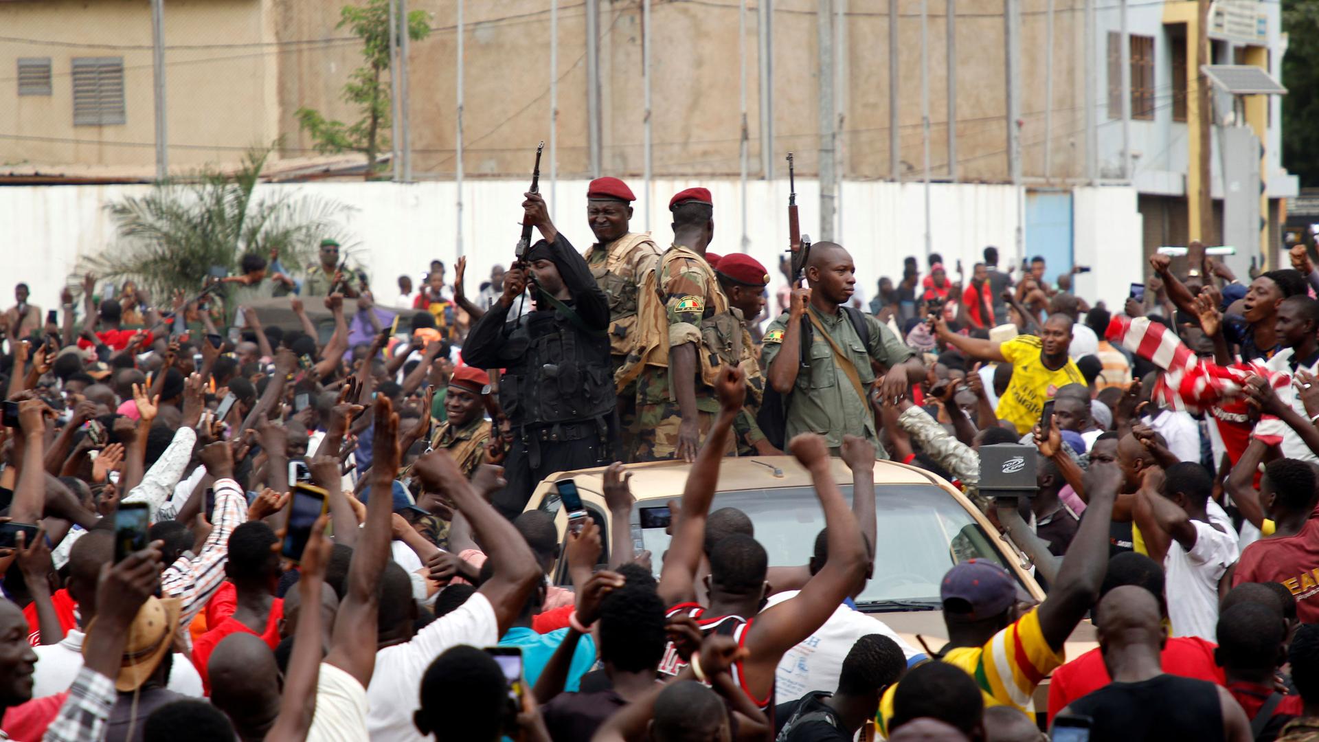 A large crowd of people are shown with their hands in the air cheering a group of armed soldiers in a car.