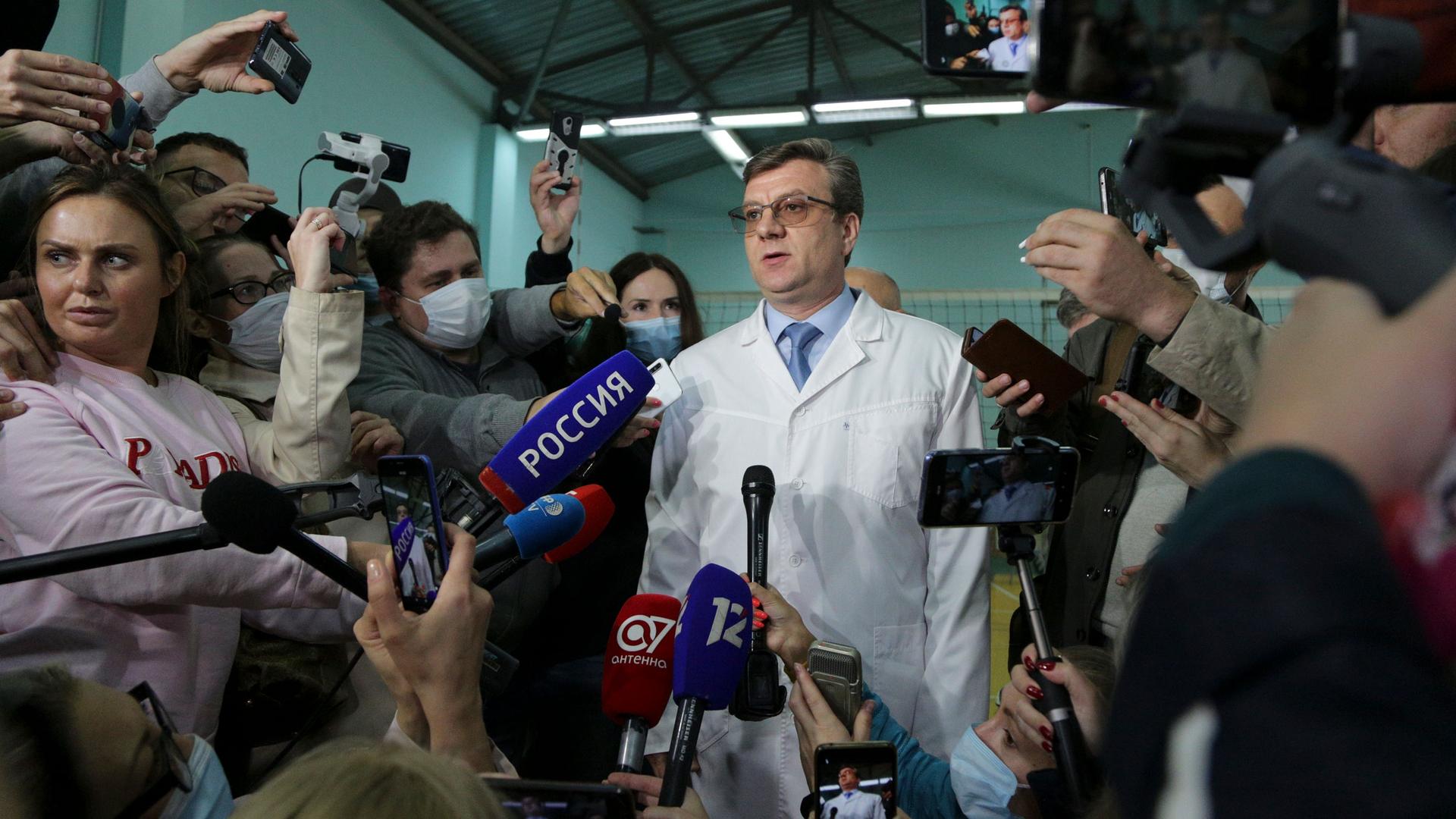 Chief physician Alexander Murakhovsky is shown wearing a white lab coat and tie while surrounded by members of the media.