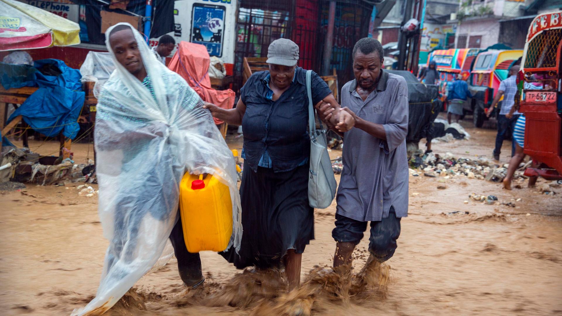 Two men are shown helping a woman as they wade through a flooded street.