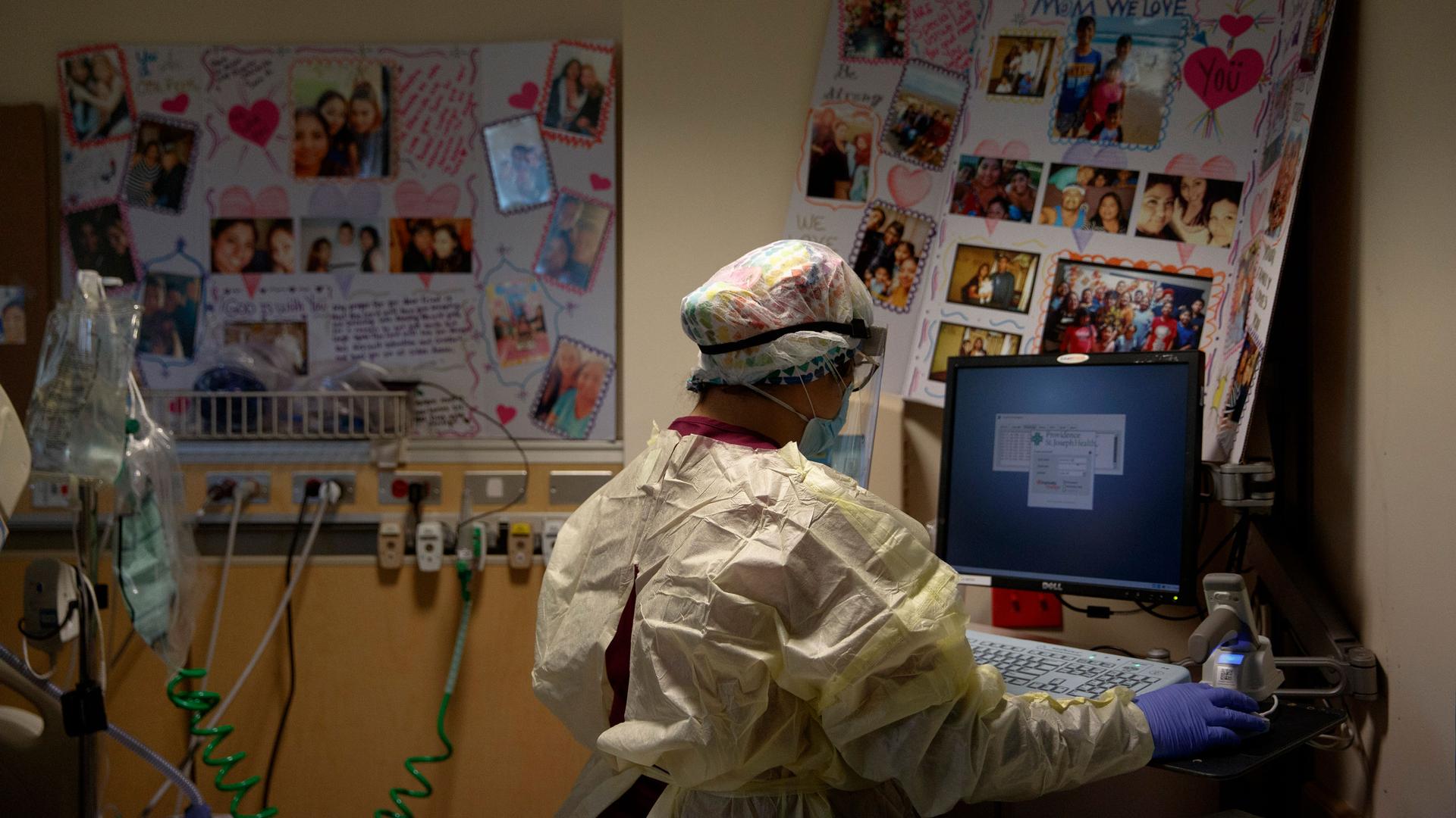 A woman is shown from behind, working on a computer and wearing a nursing cap and gown with get well soon cards on the wall.