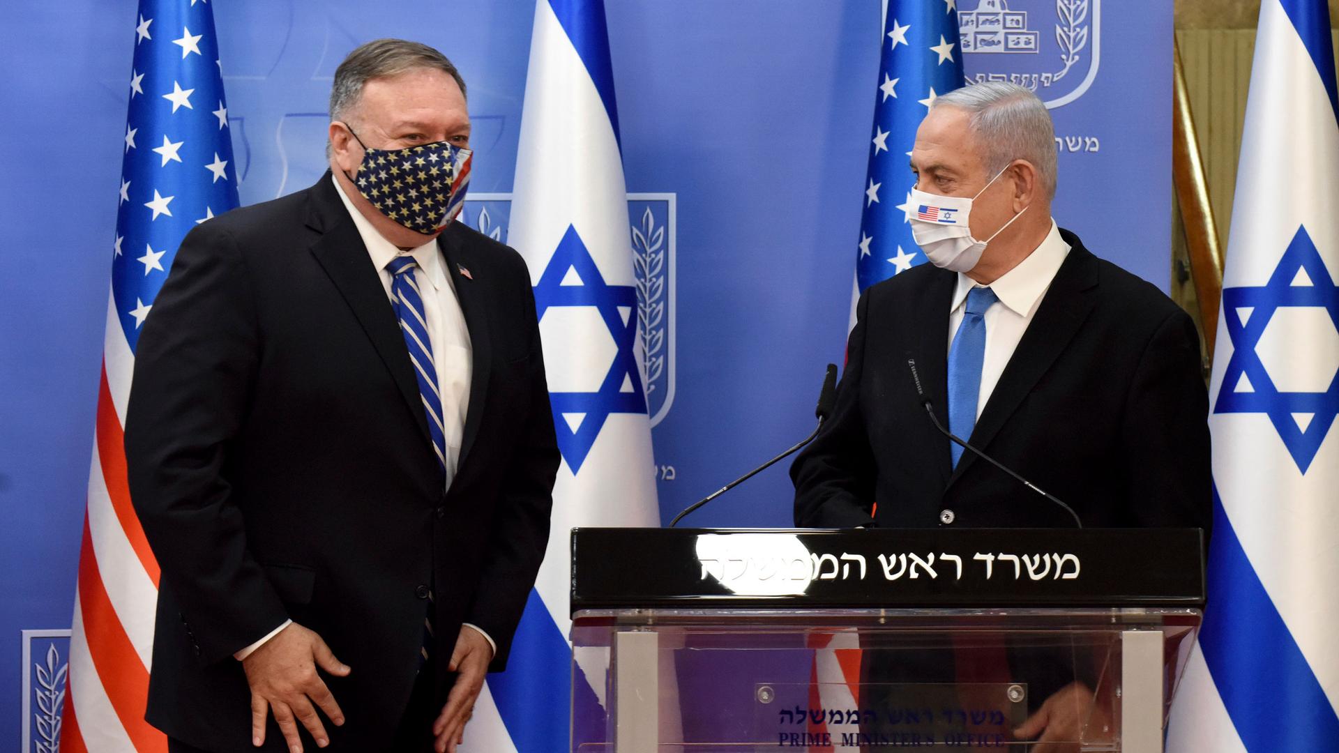US Secretary of State Mike Pompeo is shown standing next to Israeli Prime Minister Benjamin Netanyahu who is standing behind a podium, both wearing masks.