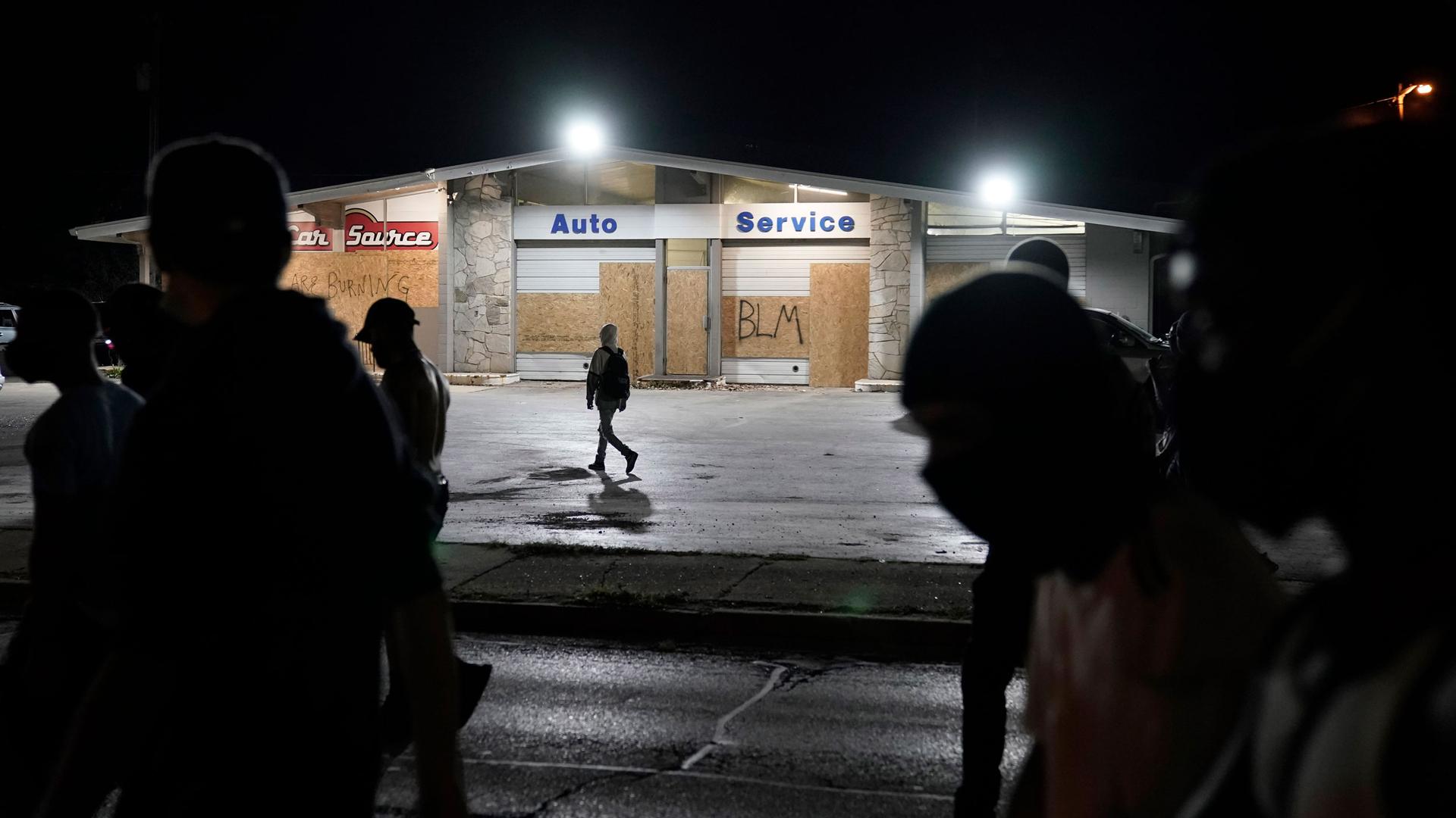 Several people are shown in shadow with a automobile service center stands in the background with boarded up windows.