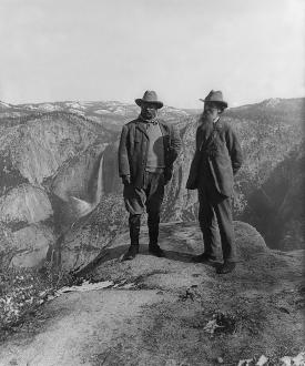 Black and white photo of two men wearing slacks, coats and hats, standing in an open landscape of rolling hills