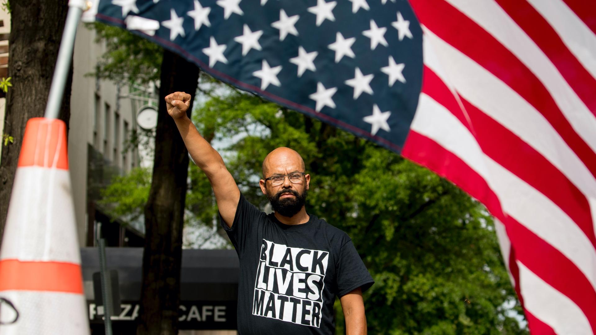 A man raises his fist and wears a black BLM shirt as he stands in the sunroof of a car with a huge American flag. 