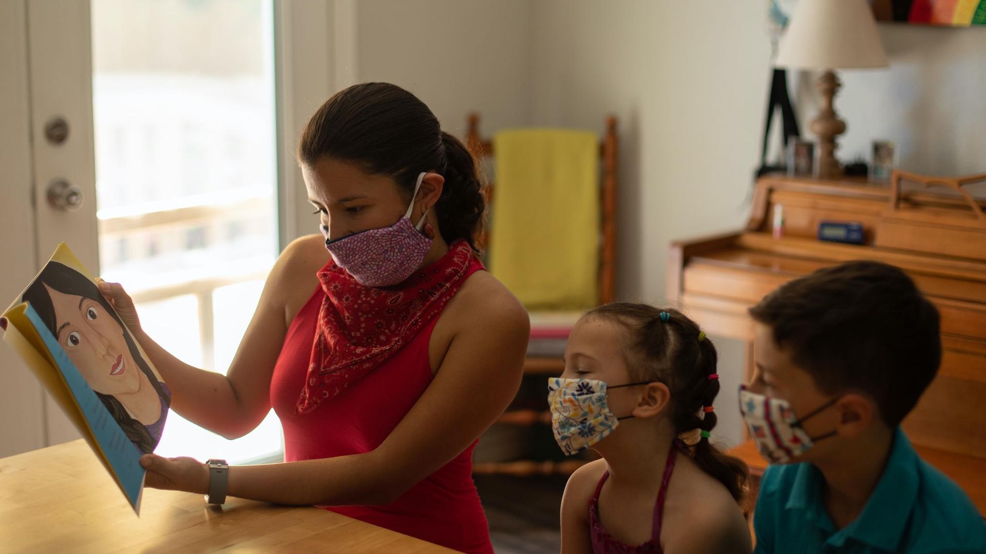A woman wears a mask and reads a book to two children, also wearing masks. 