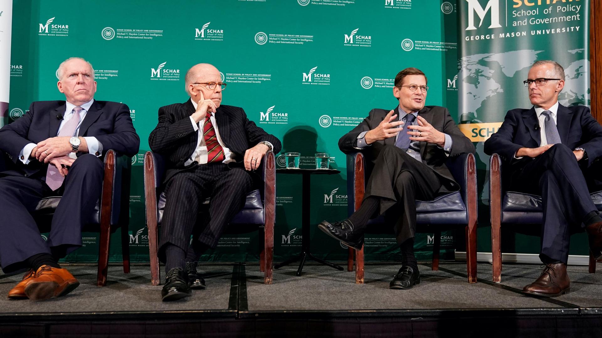 Four white men in suits sit on a panel with a green background