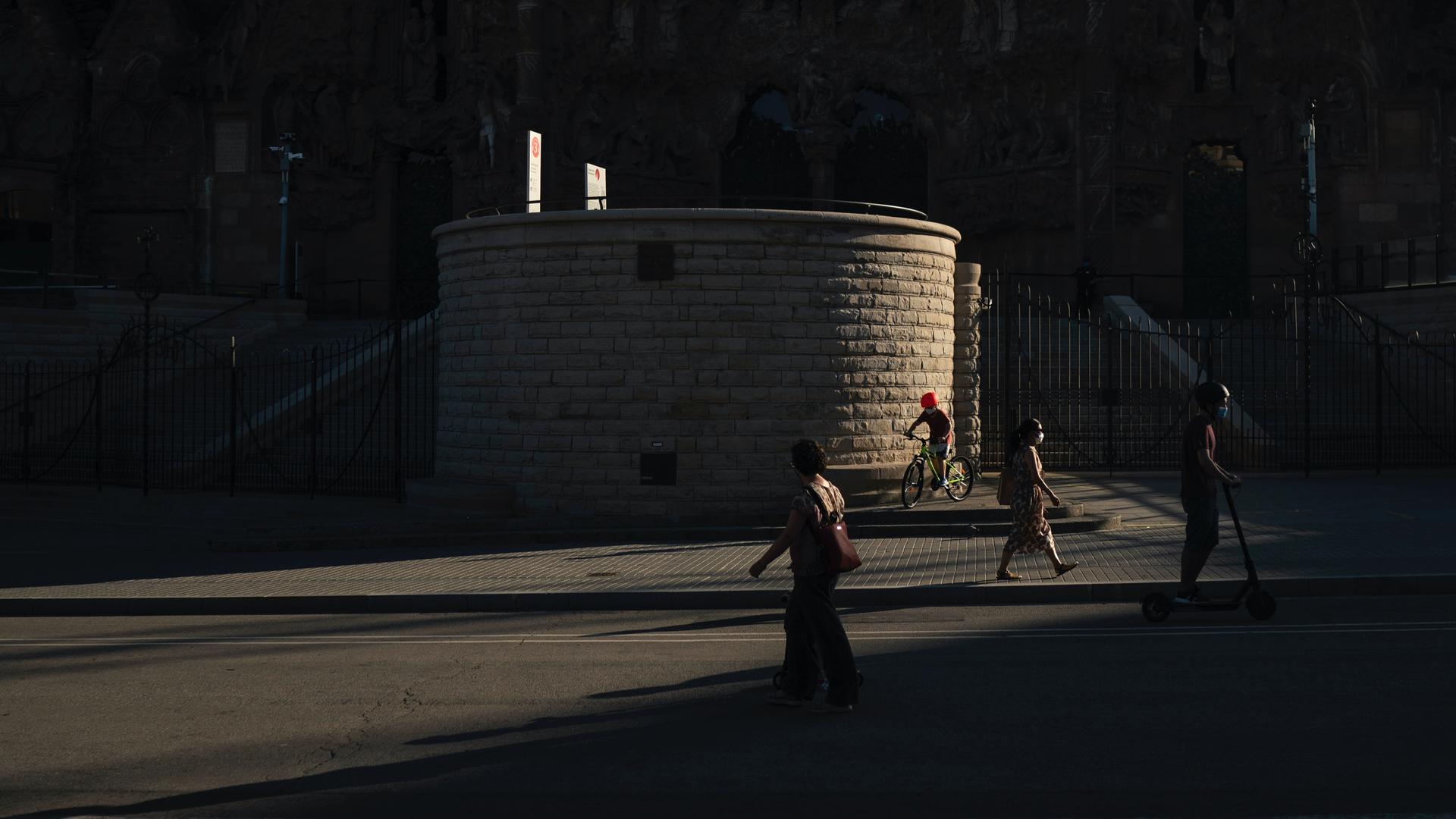A few people walk or bike in shadows near a large building. 