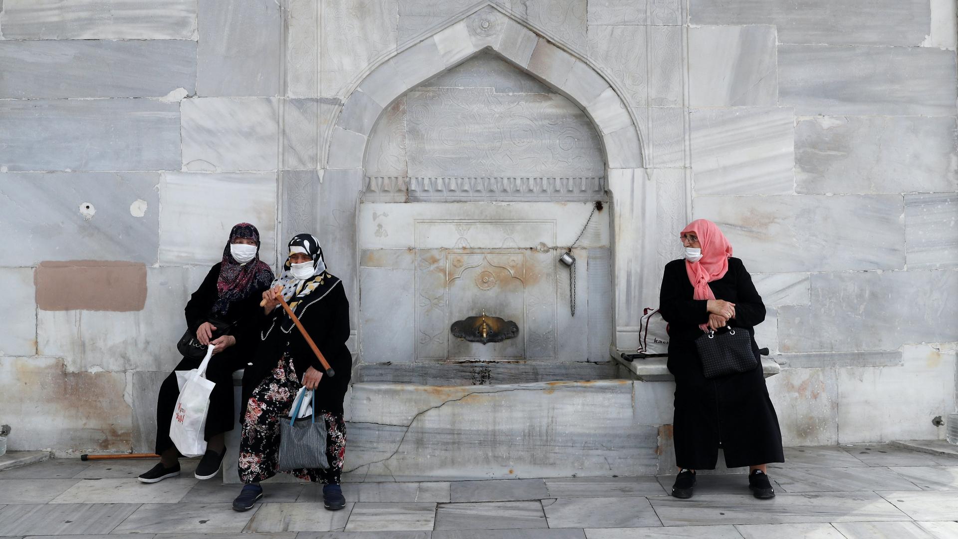 Three women wearing head scarves, long jackets and masks by a gray fountain 