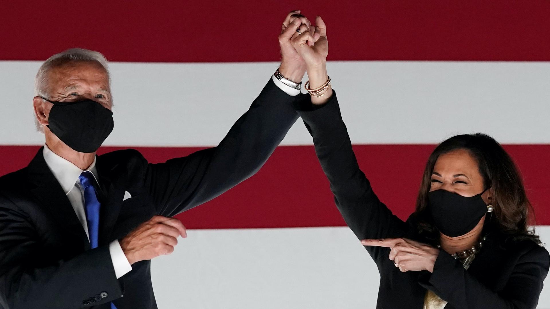 Biden and Harris wear masks, clasp hands, and Harris points at Biden, smiling with her eyes. 