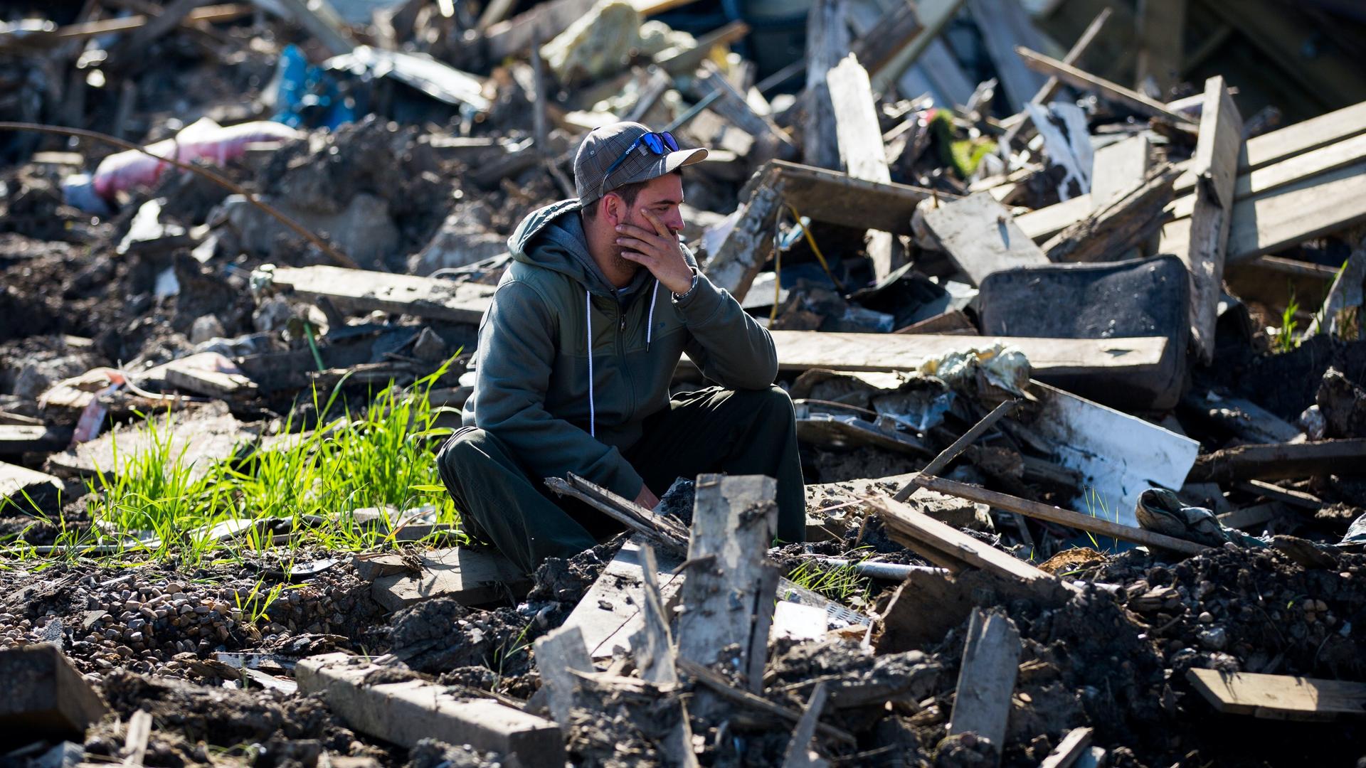 A man squats in rubble holding his head in a perplexed way