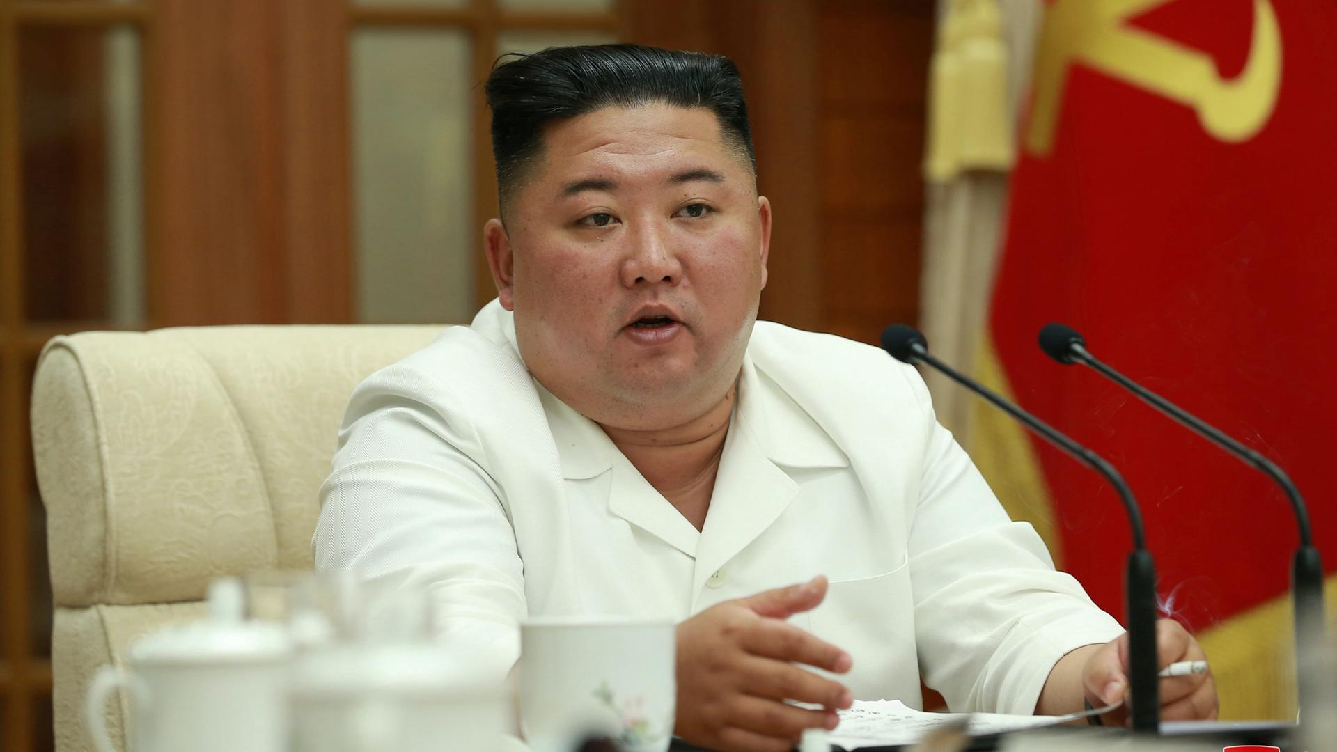 North Korean leader Kim Jong-un, with a white suit and black flat-top hair style, speaks at a conference table. 