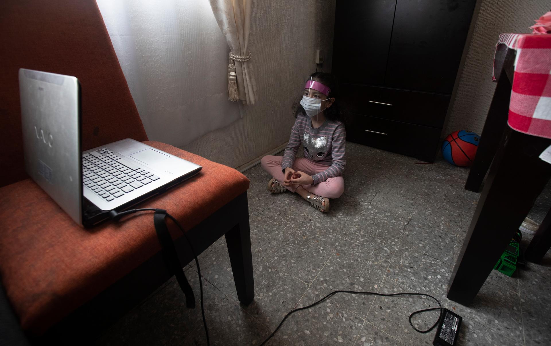 A girl wears a plastic guard indoors as she watches a laptop screen.