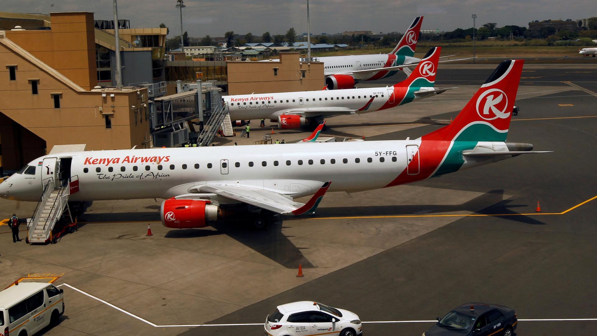 Several white airplanes with Kenya Airways logo on them in red and green parked in airport. 