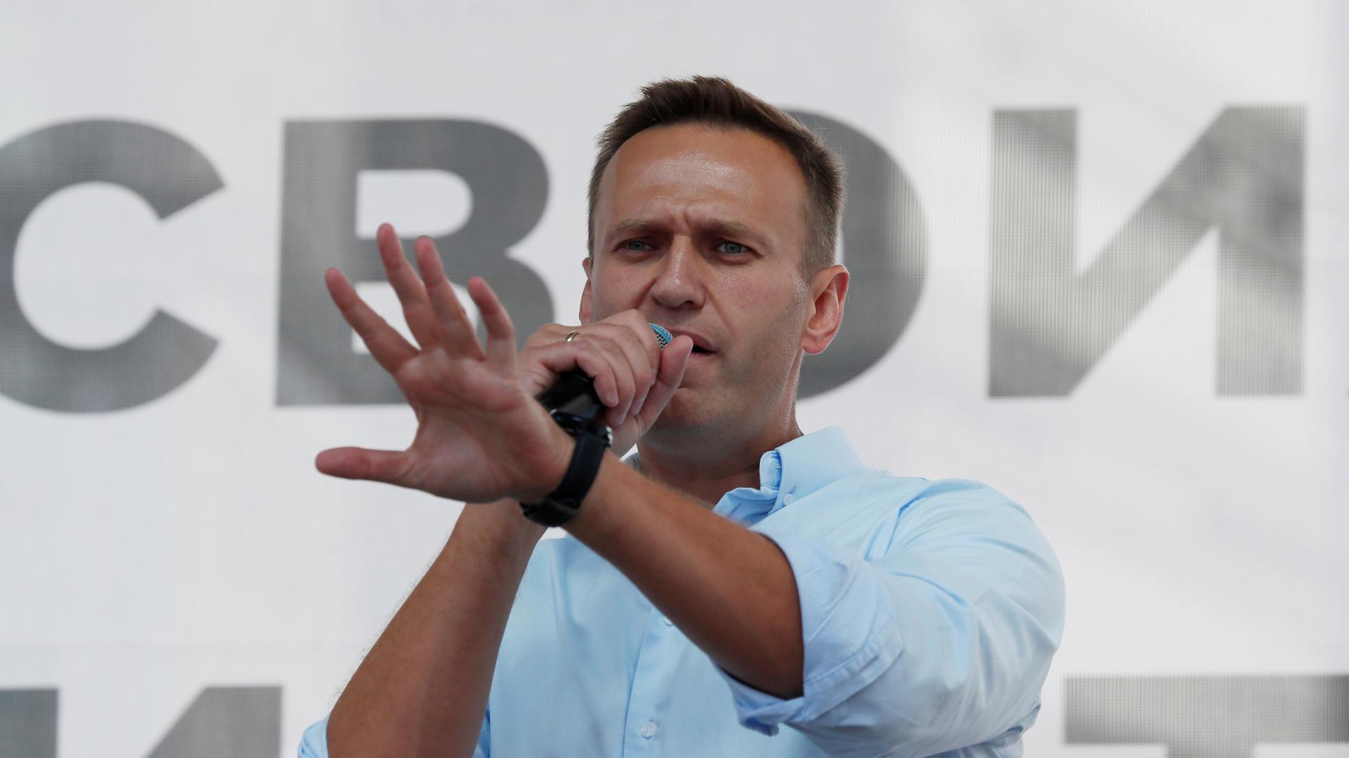 A man wearing a white shirt speaks on a microphone while gesturing with his hands.