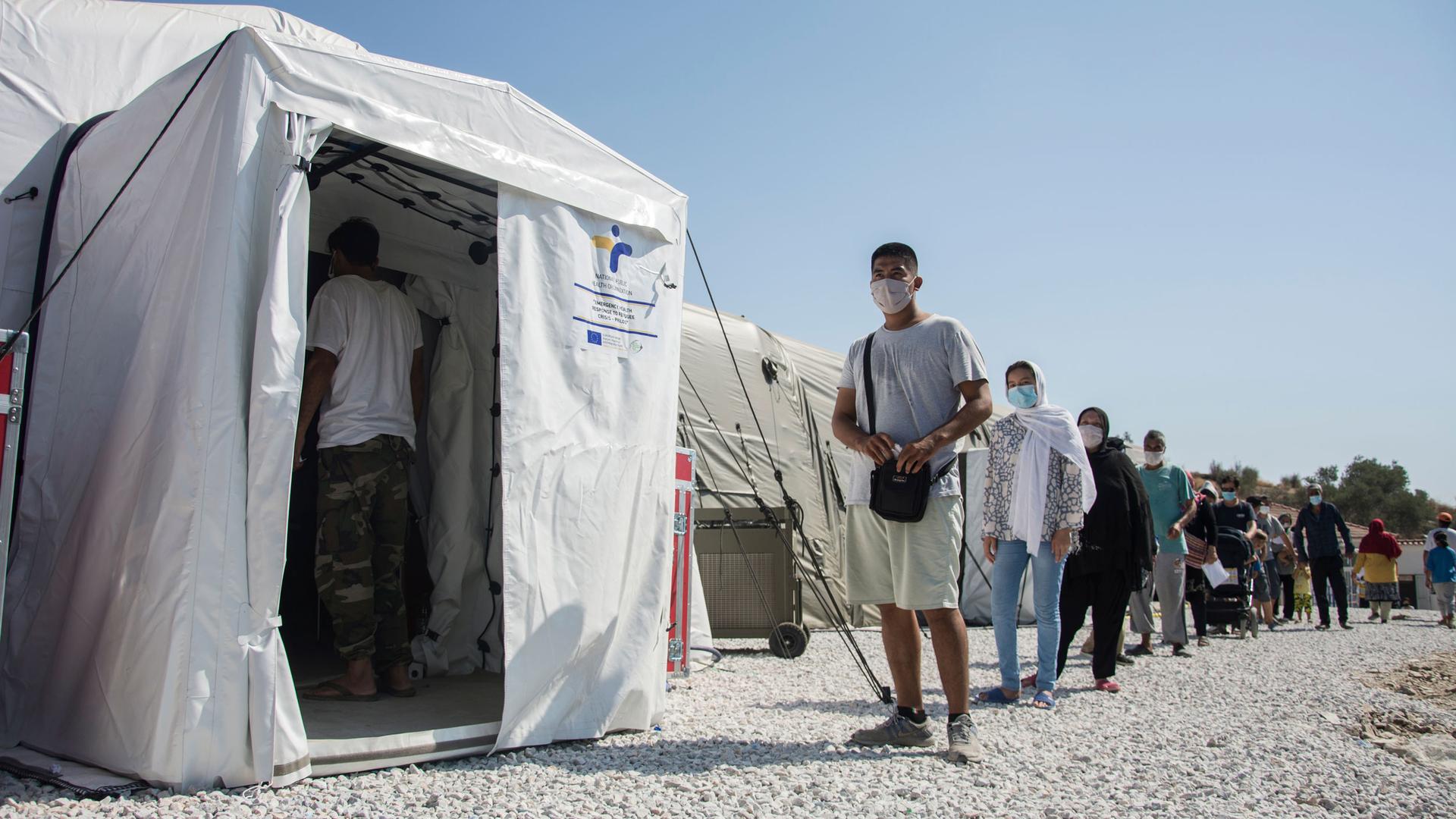 A white medical tent is shown with a long line of people waiting outside of it.