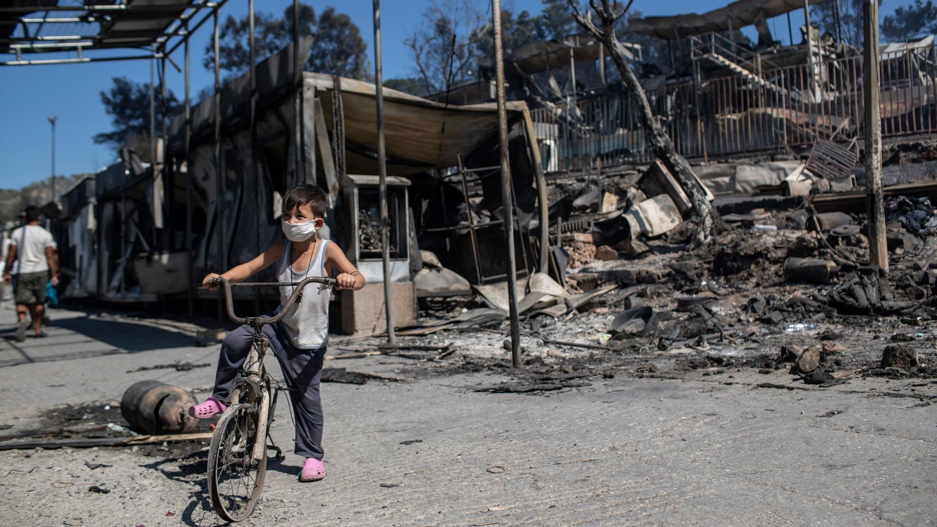 A young boy is shown sitting on a bike without tires in front of a destroyed structure at a refugee camp.