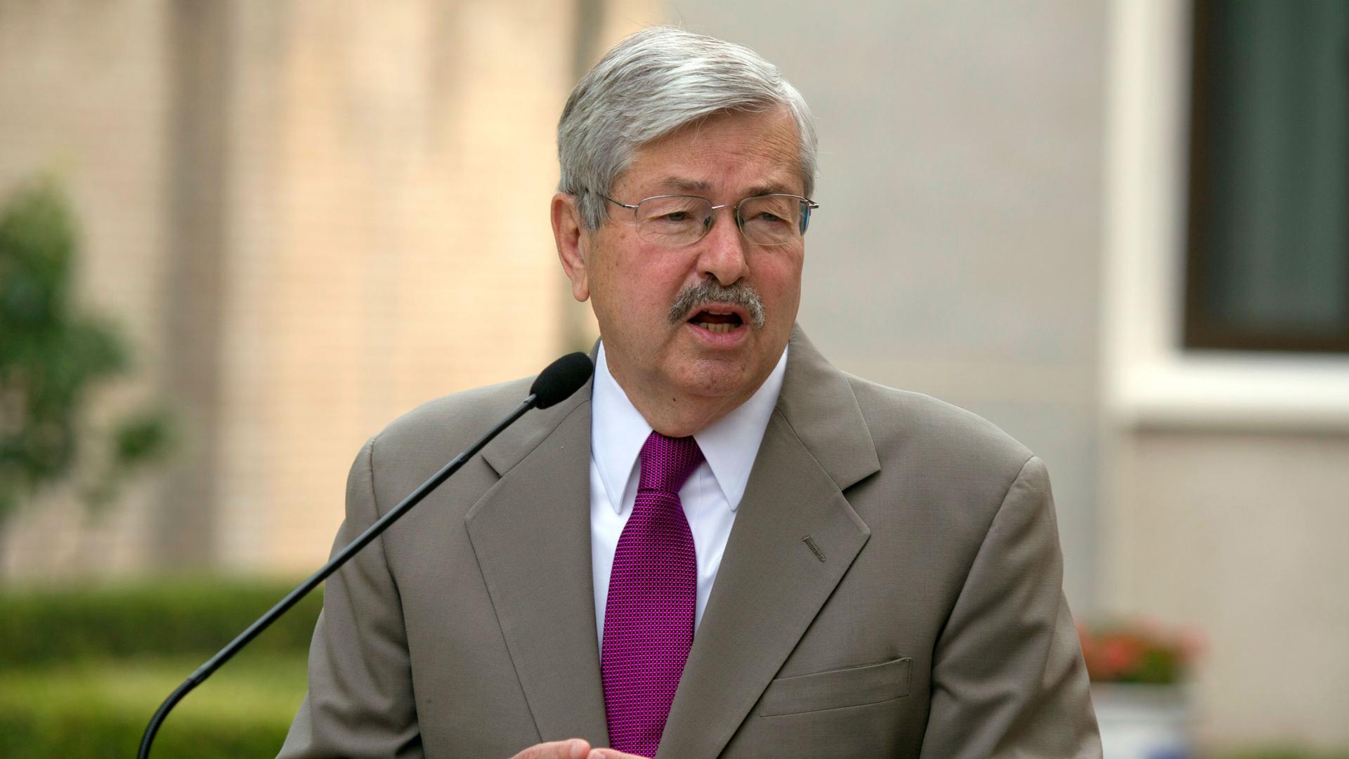 US Ambassador to China Terry Branstad is shown wearing a tan suit and a purple tie while speaking into a small microphone.