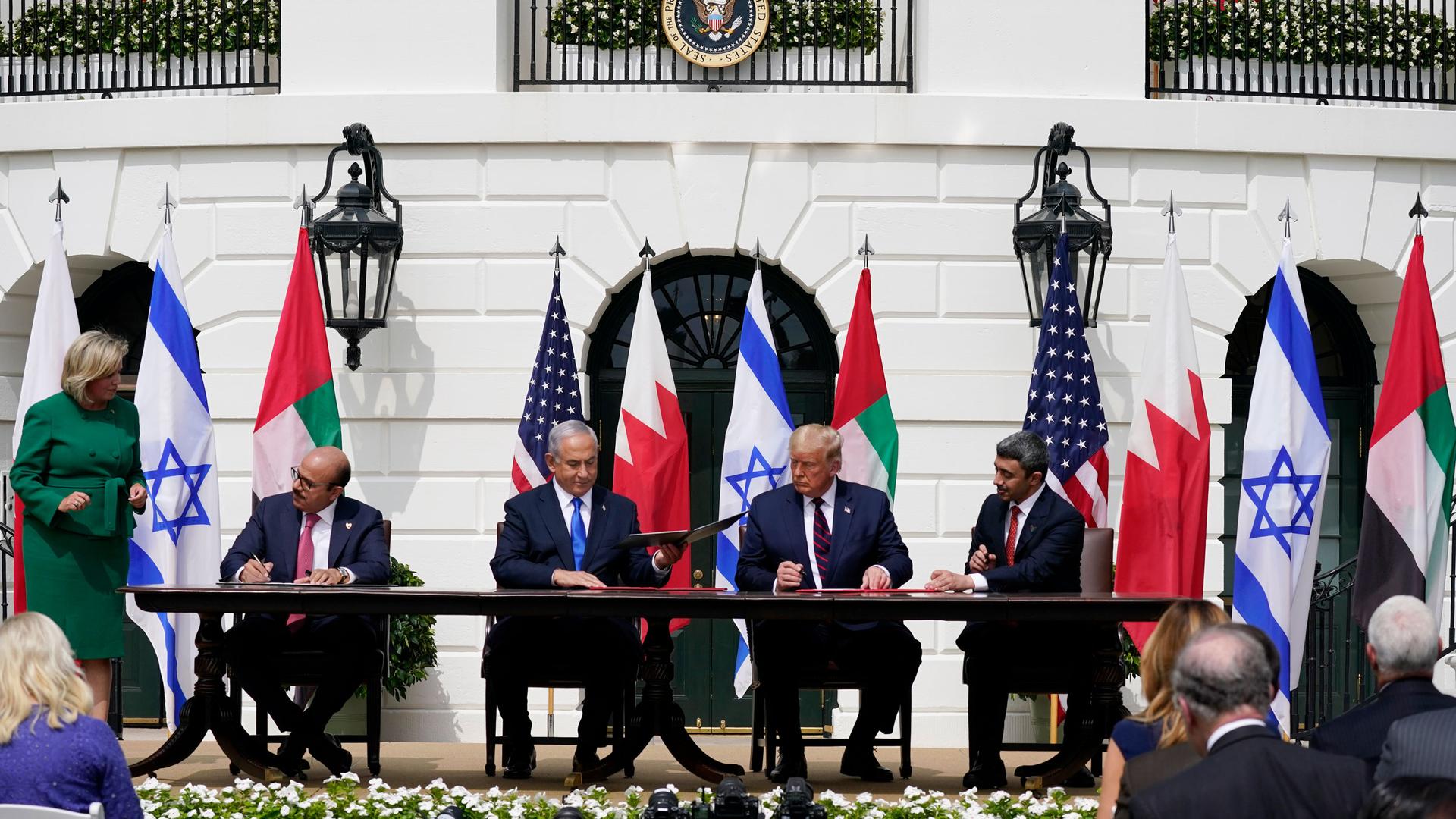 The leaders of the US, UAE, Israel and Bahrain are shown wearing suits and sitting at a table signing documents with flags from each country behind them.