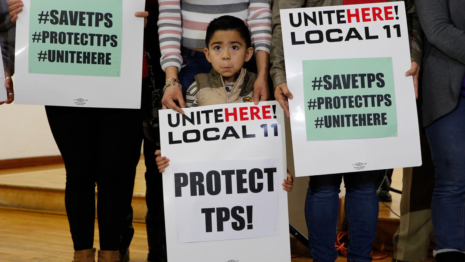A young boy is shown holding a large white sign that says, "Unite here! Local 11, Protect TPS!" on it.
