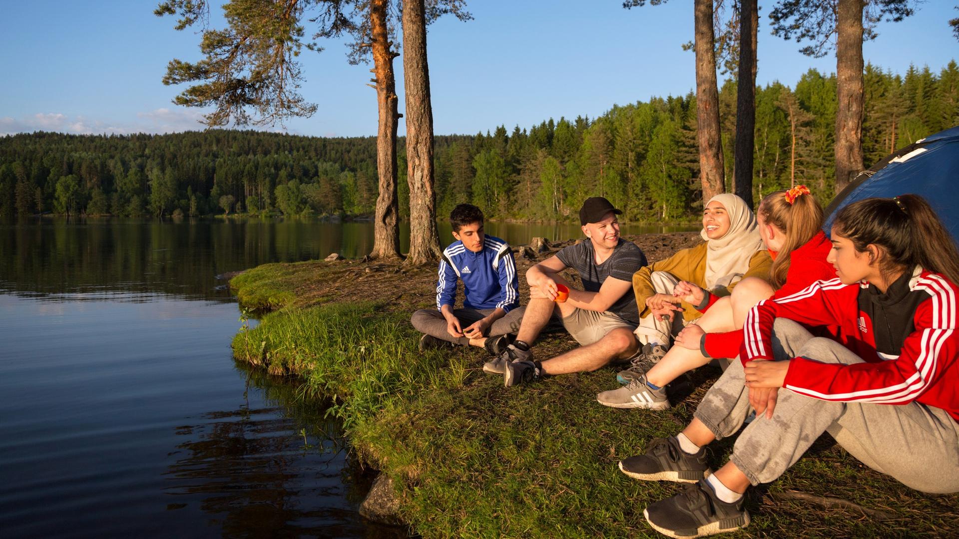Norwegian youth sit on the ground while spending time outdoors on a lake.