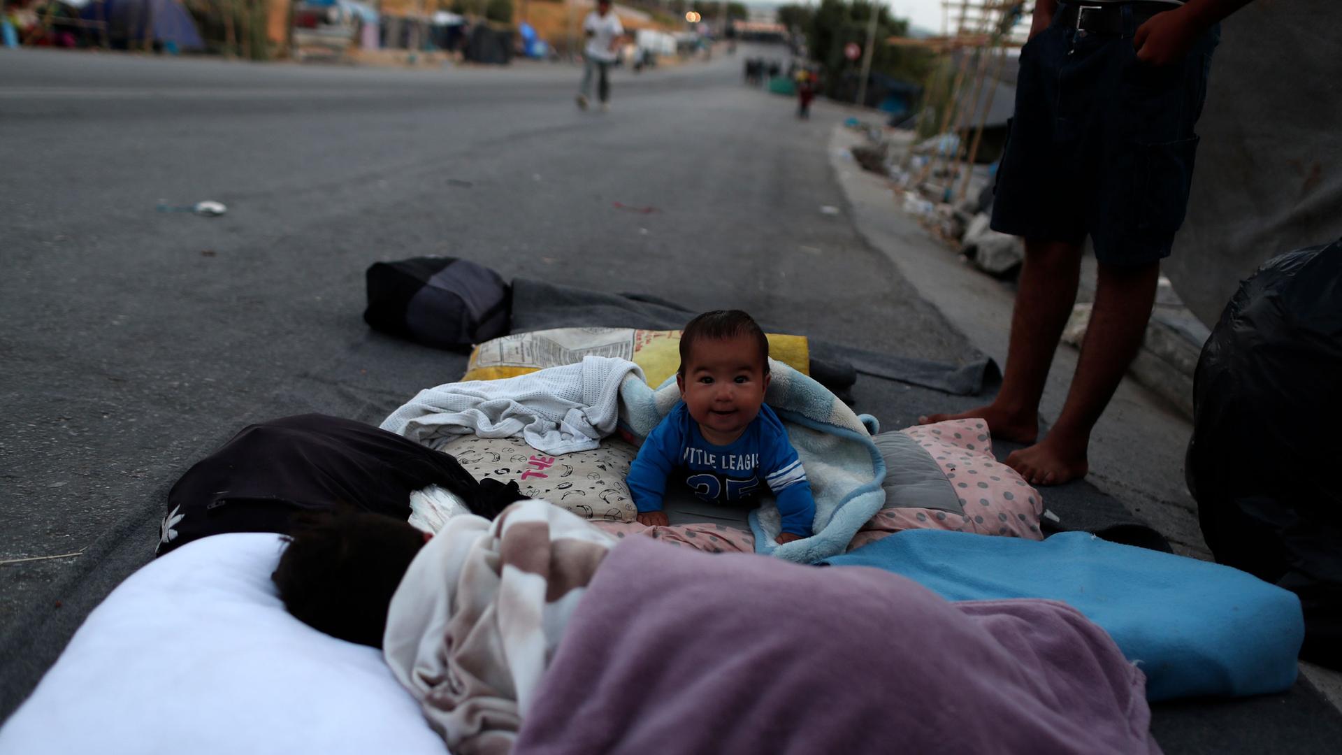 A small baby is shown looking at the camera and crawling on blankets out on a street.