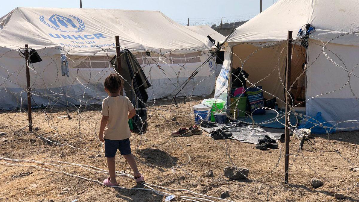 A young child is shown wearing shorts and a t-shirt walking past a barbed-wire fence and white UNHCR tents.