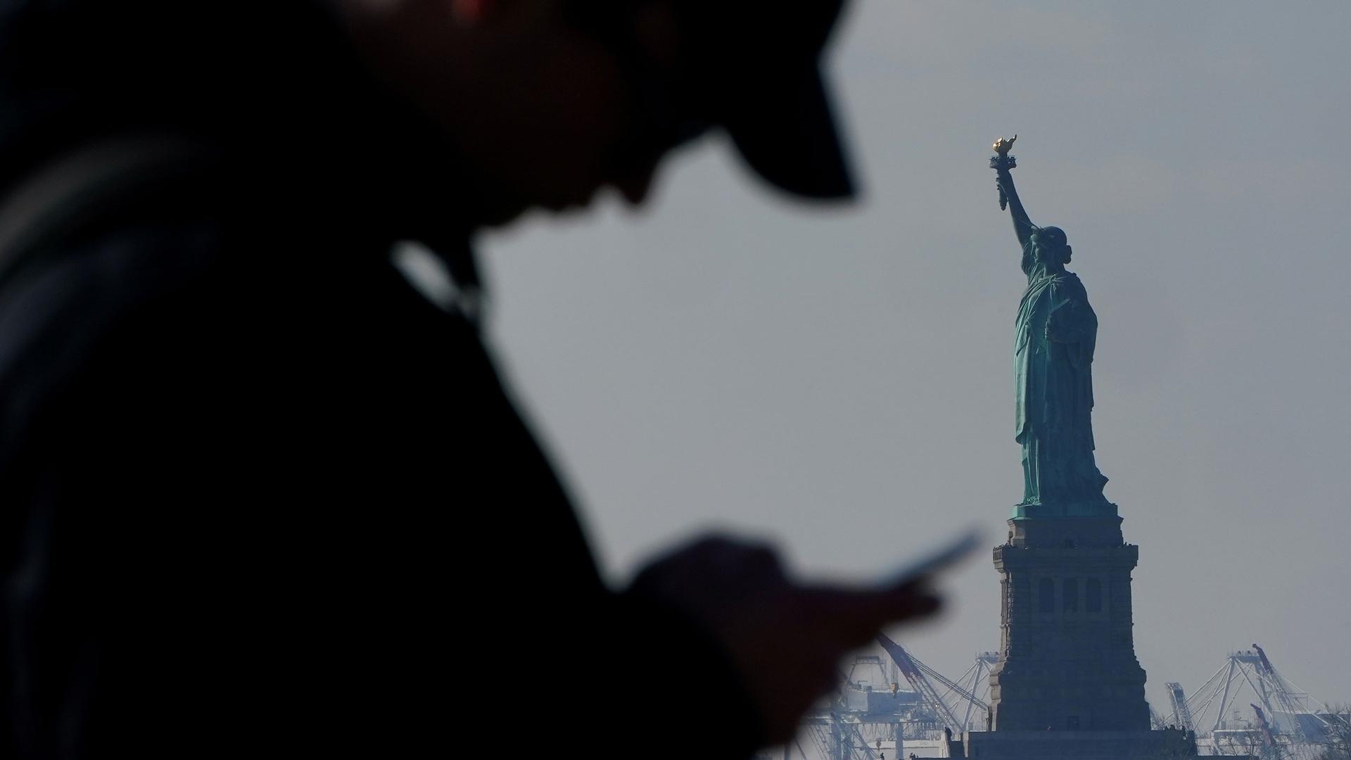 A man is shown in shadown hearing a hat and looking at a mobile phone with the Statue of Liberty in the background.
