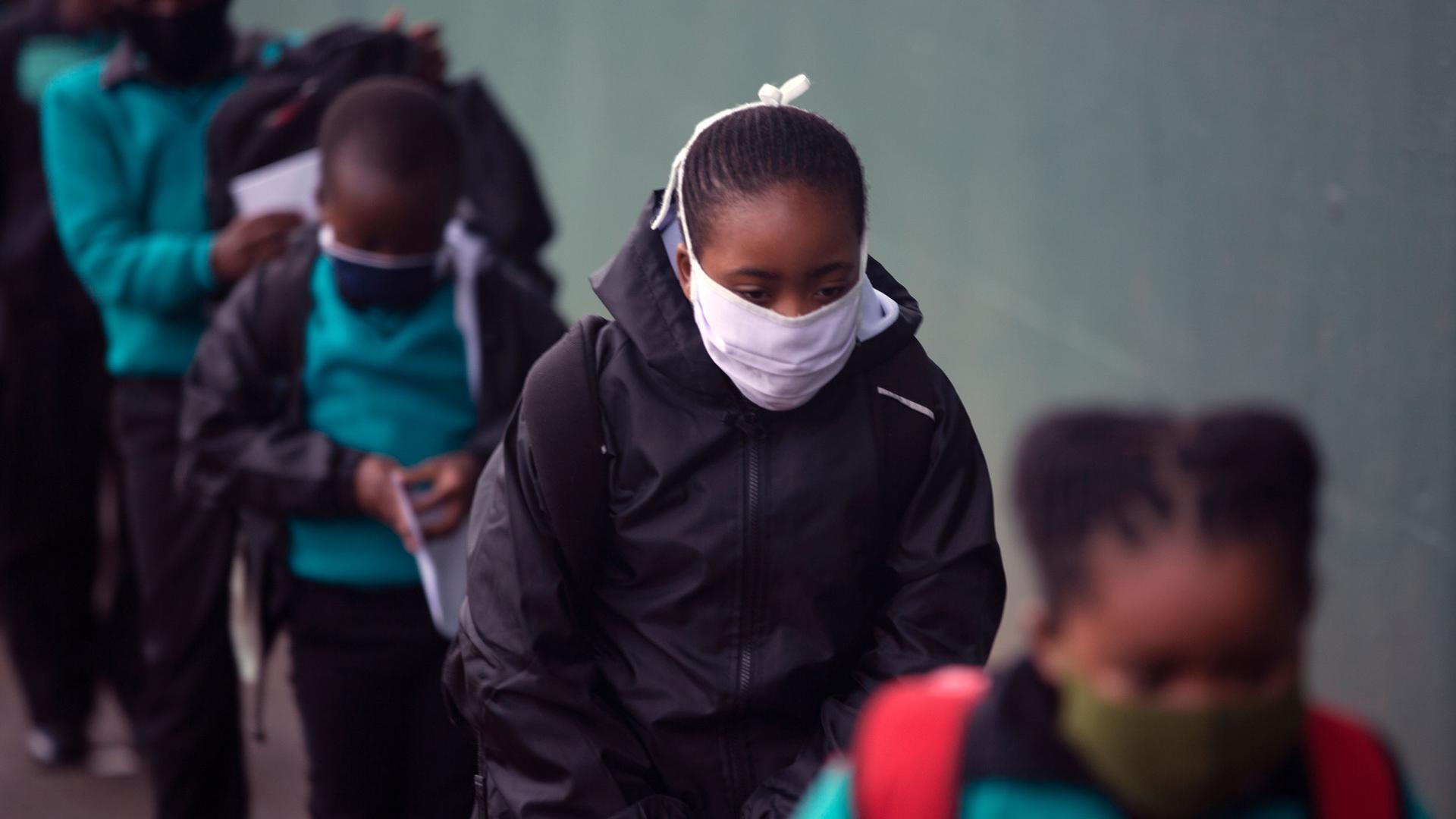 A line of young school children are shown wearing face masks and wearing backpacks and jackets.