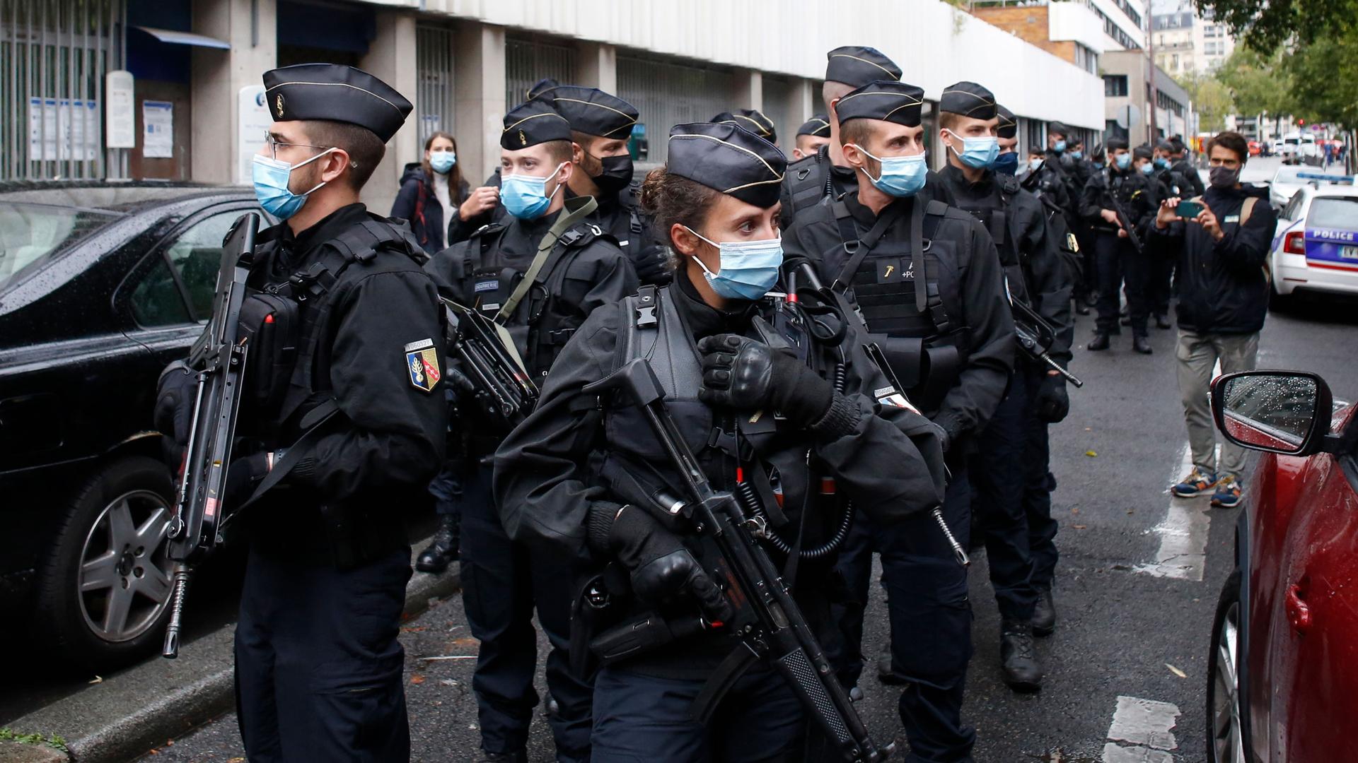 A group of police officers are shown carrying large weapons and wearing face masks while walking down the middle of a street.